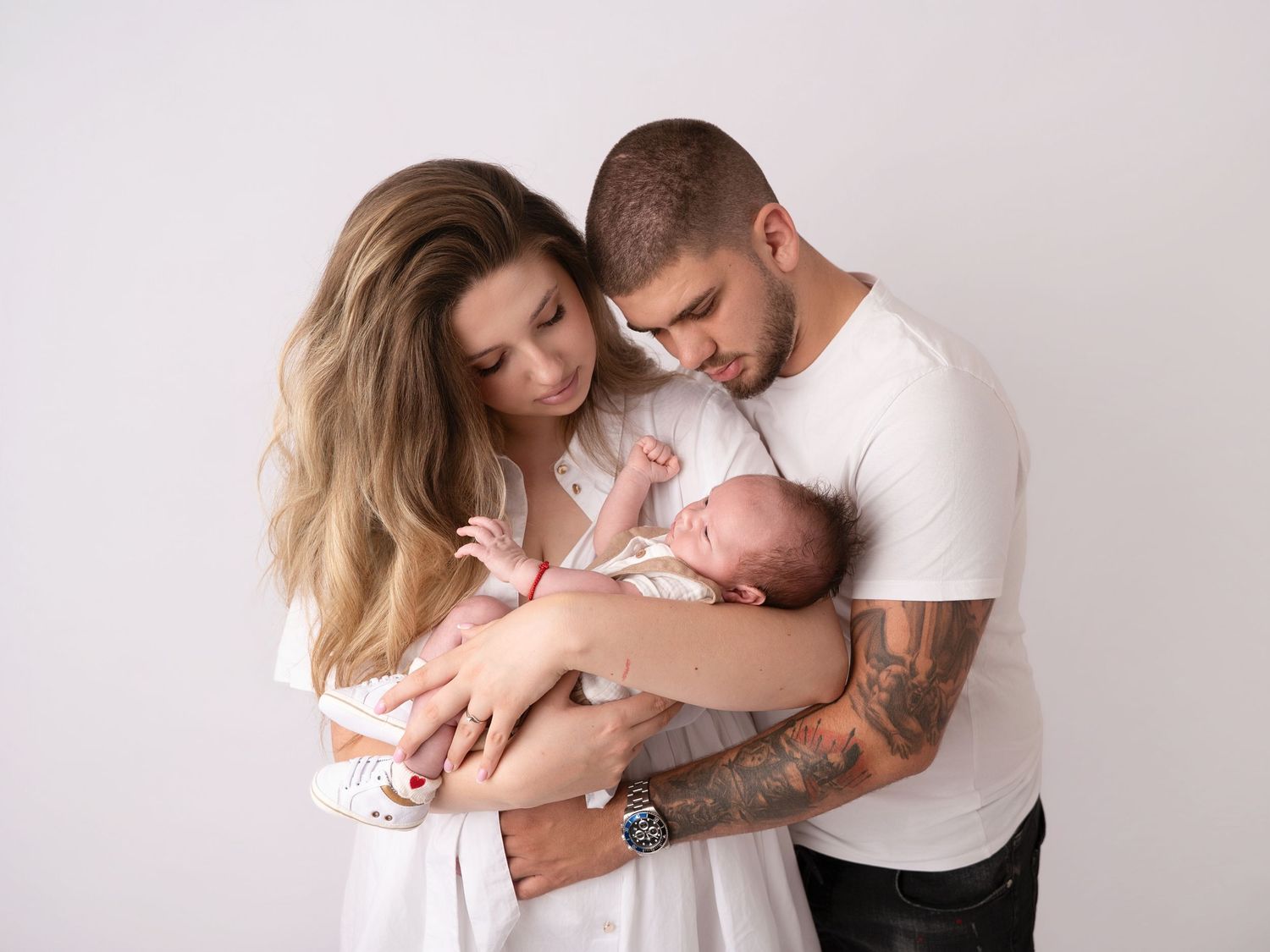 A young couple in white outfits tenderly holds their newborn baby together against a soft white studio background.