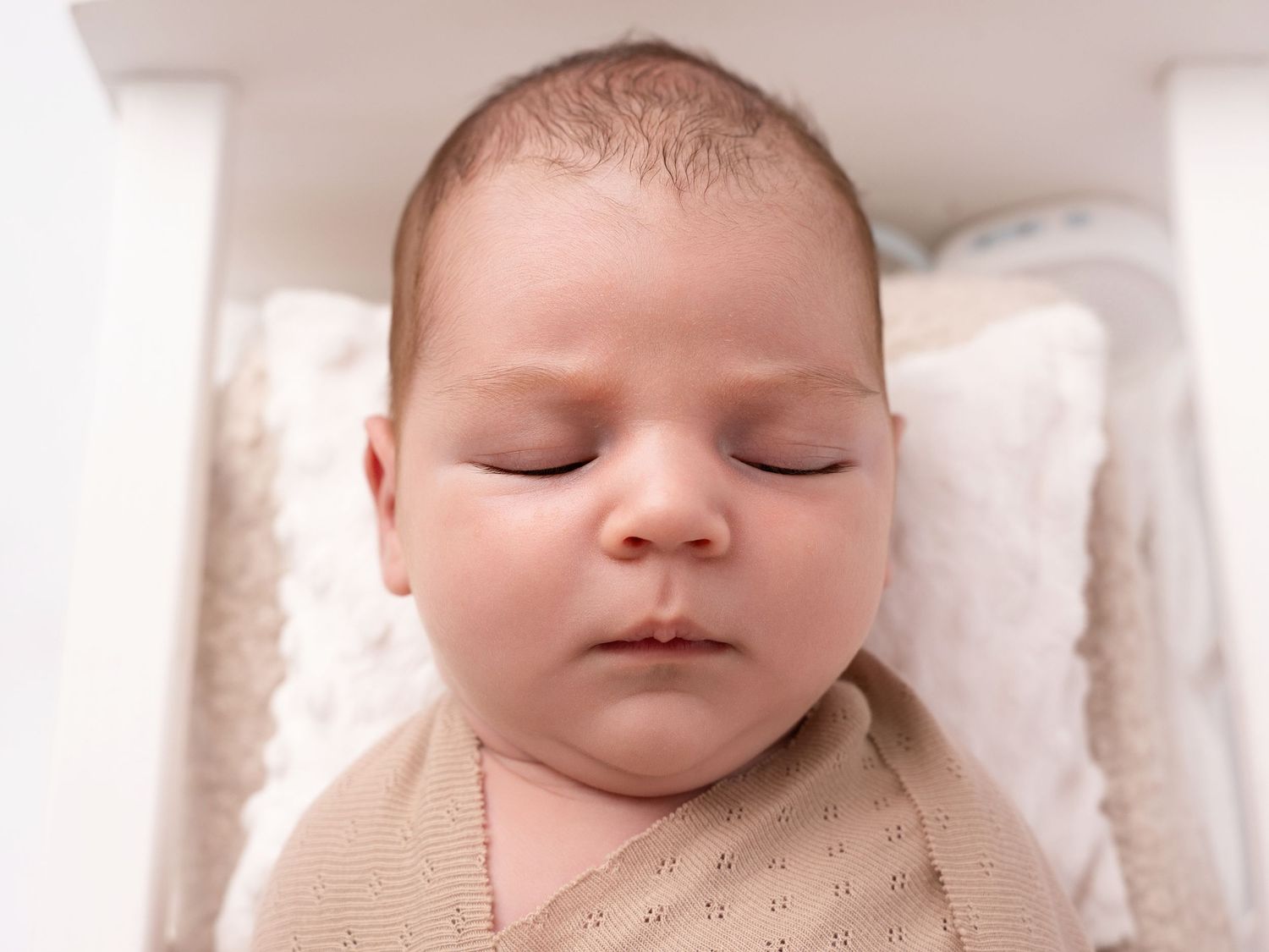 Peaceful sleeping newborn baby swaddled in a soft beige knit blanket, lying in a white crib.