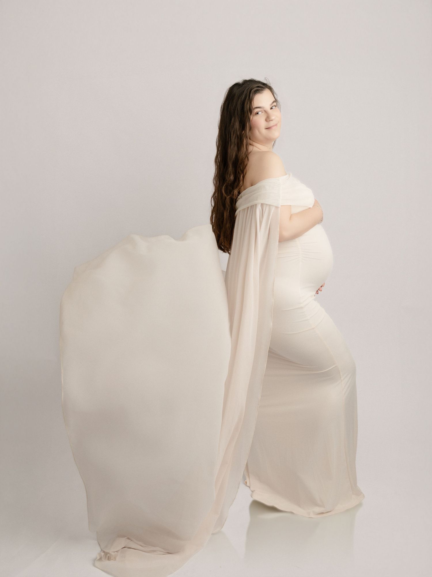 Pregnant woman in white off-shoulder maternity gown with flowing fabric cape posing sideways in bright studio.