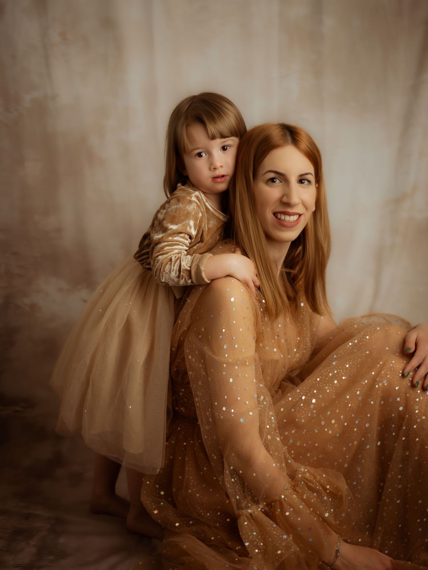 A smiling red-haired woman and young girl in matching gold sparkle dresses pose together in a studio.