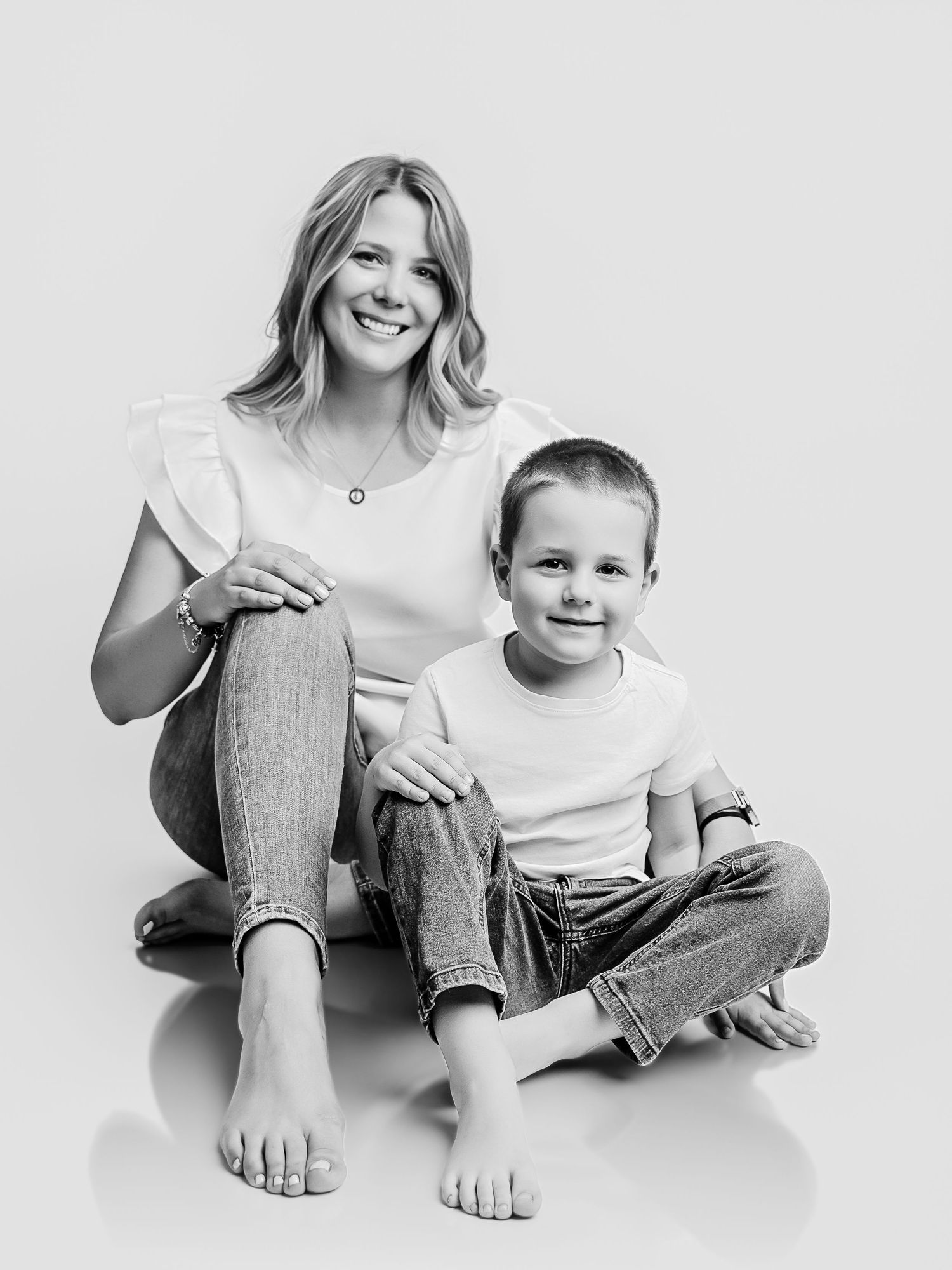 Black and white studio portrait of a smiling mother and young son sitting together on a white background.