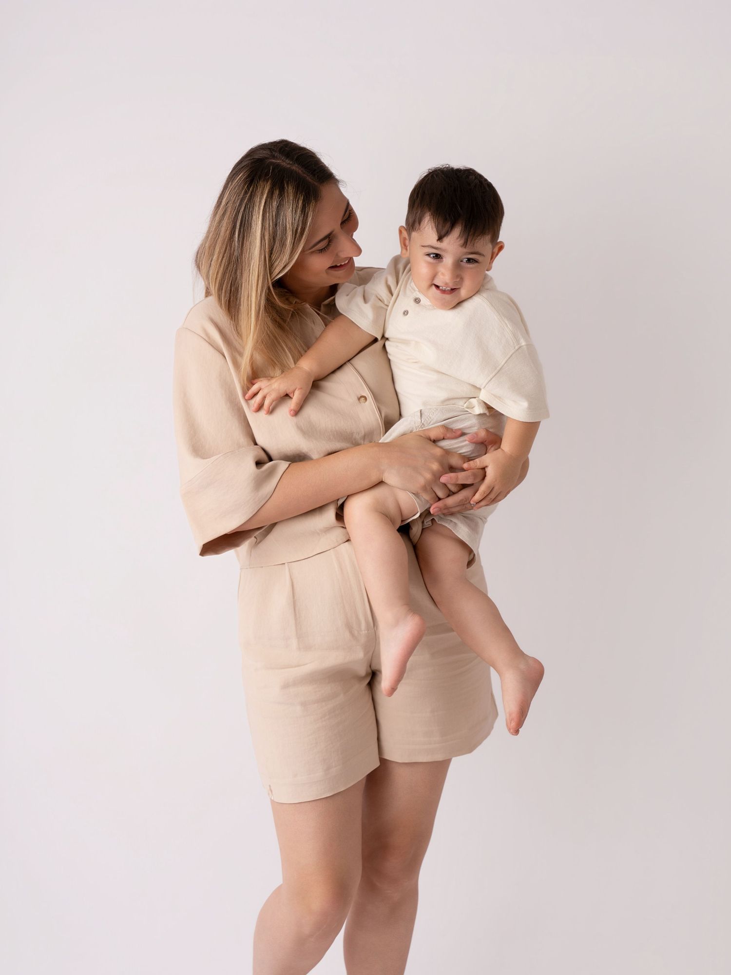 Woman in beige linen shorts set holding a smiling toddler boy in a cream outfit against a white background.