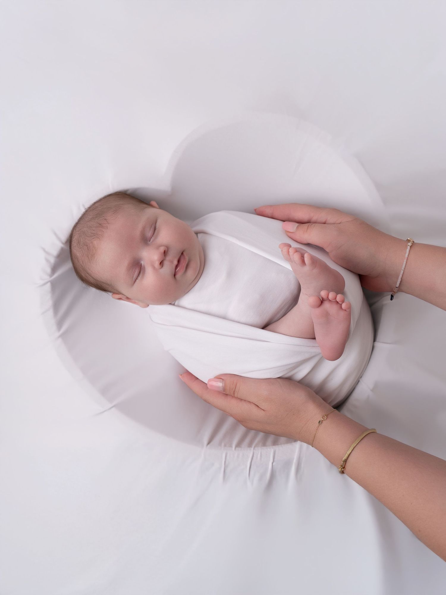 Sleeping newborn baby in white swaddle rests on soft white pillow while mother holds baby gently.