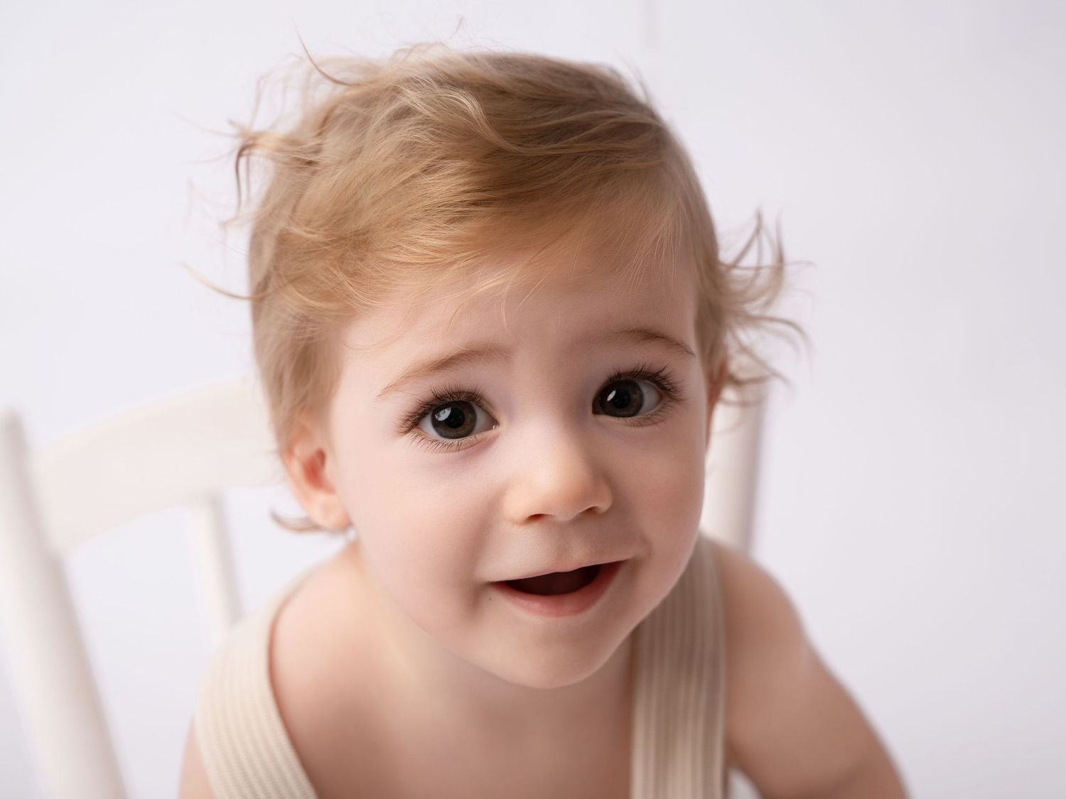 Adorable toddler with light brown hair and big brown eyes sitting in a white chair against a white background.