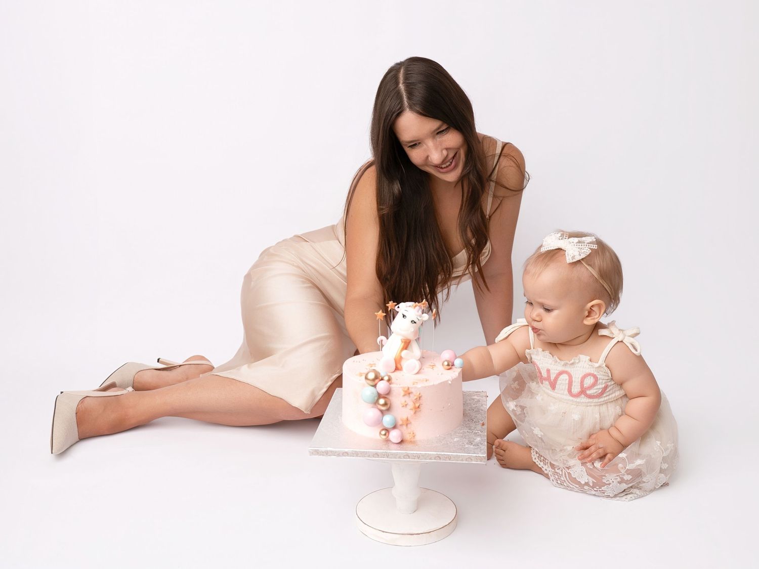 A mother in a beige dress helps her baby girl touch a pink first birthday cake during a studio smash cake session.
