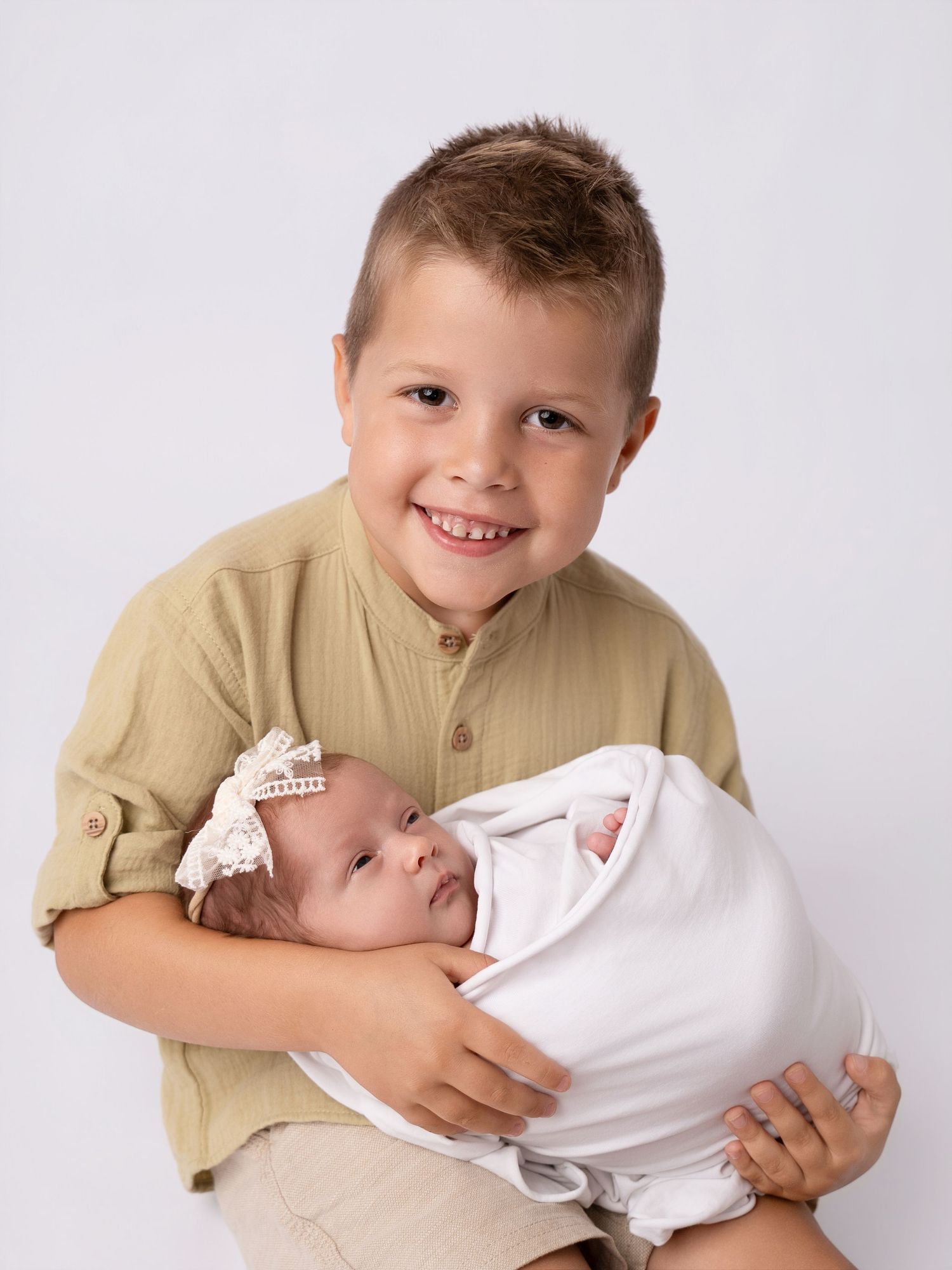 Smiling young boy holding a swaddled newborn baby girl with a white bow, professional sibling portrait photo.