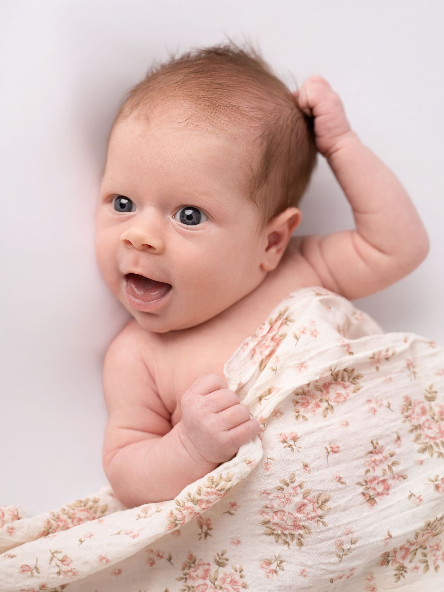 Adorable newborn baby with blue eyes and reddish hair, wrapped in a floral muslin swaddle, smiling on a white background.