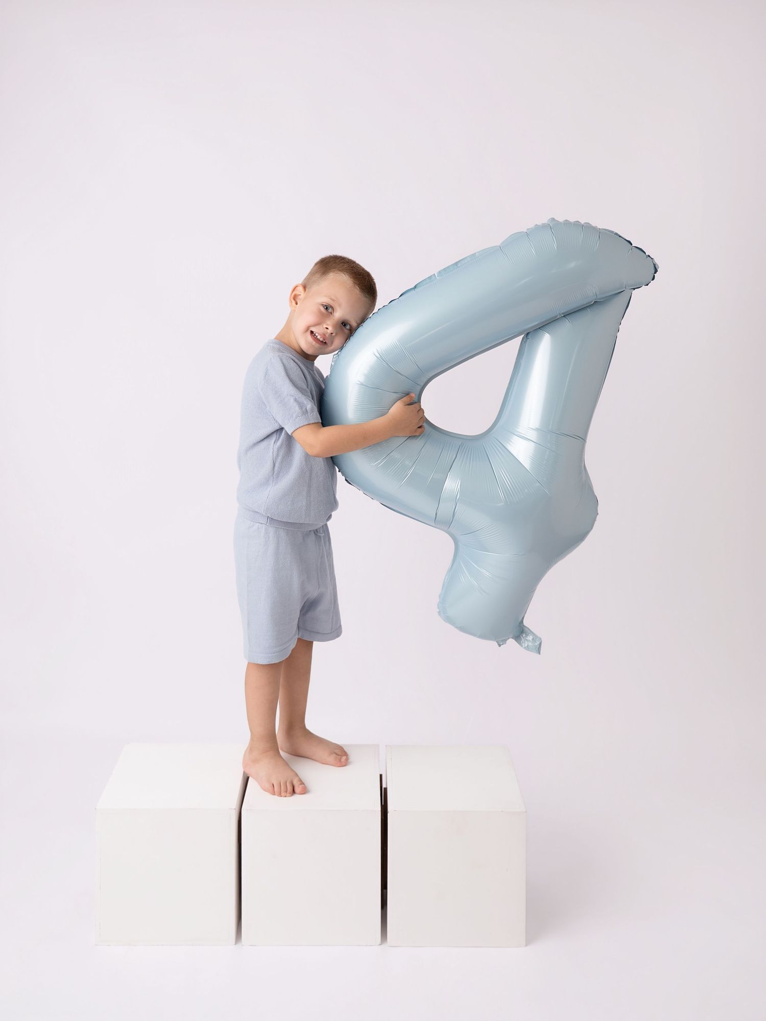 A smiling young boy stands on white boxes holding a large light blue number 4 foil balloon against a white background.
