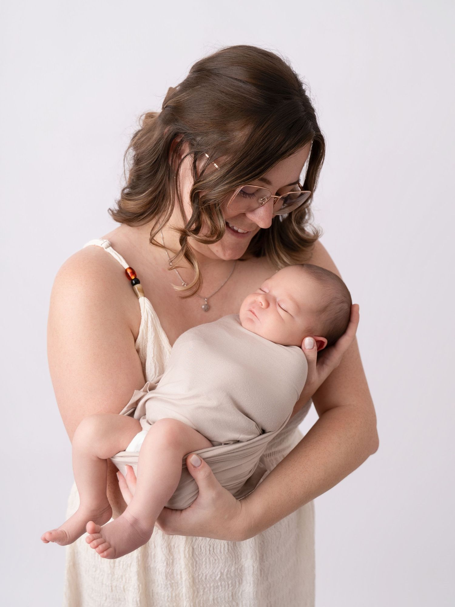 A smiling mother with glasses tenderly holds her sleeping newborn baby wrapped in a beige swaddle.