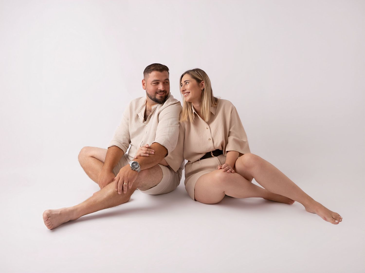 A couple in matching beige outfits sit back-to-back on a white studio floor, smiling warmly at each other.