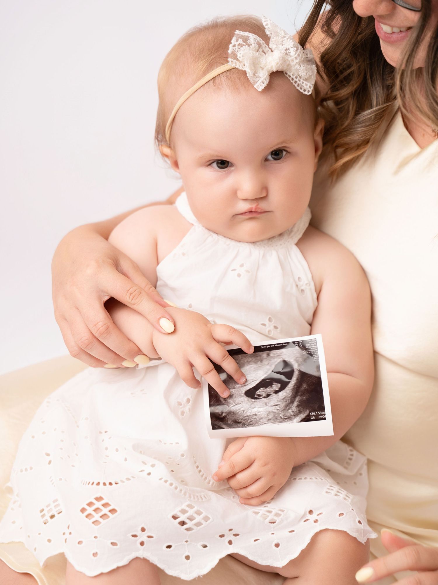 A baby girl in a white dress holds an ultrasound photo while sitting with her mother during a maternity announcement shoot.