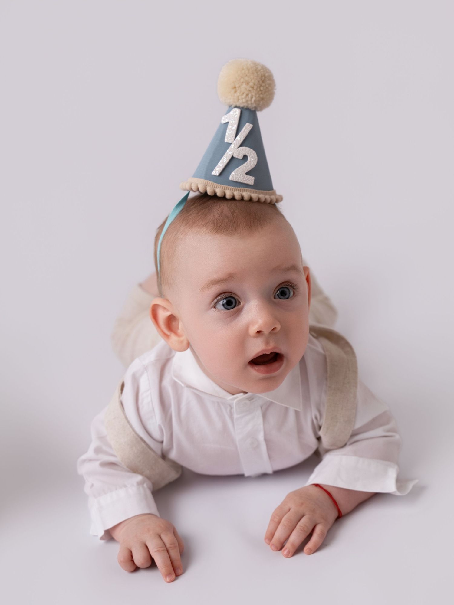 Adorable baby wearing a blue half birthday party hat with pom pom, lying on tummy on white background.