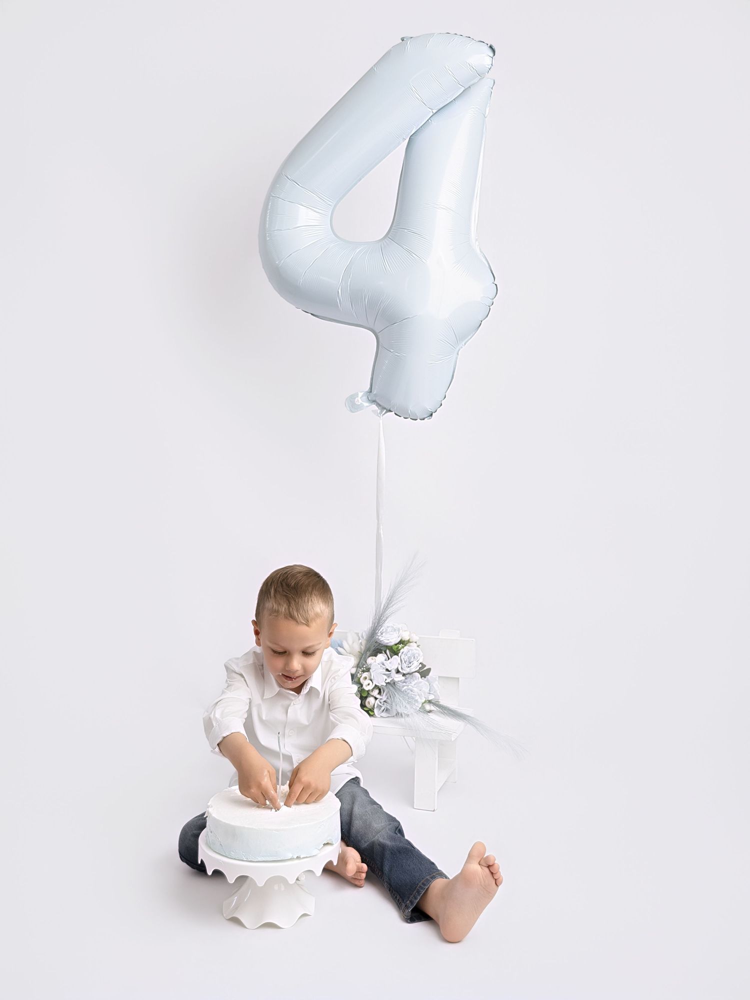 A young boy sits on the floor with a white cake, next to a large light blue number 4 balloon for his birthday.