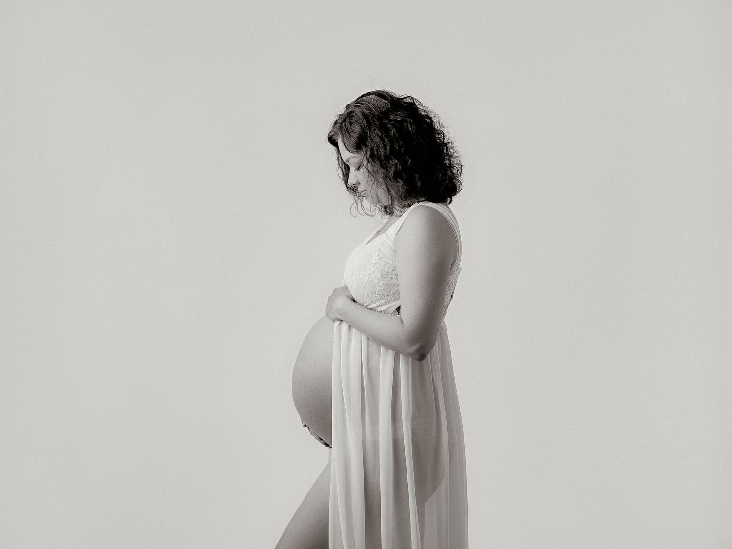 Black and white maternity photo of pregnant woman in white dress gently holding her bump.