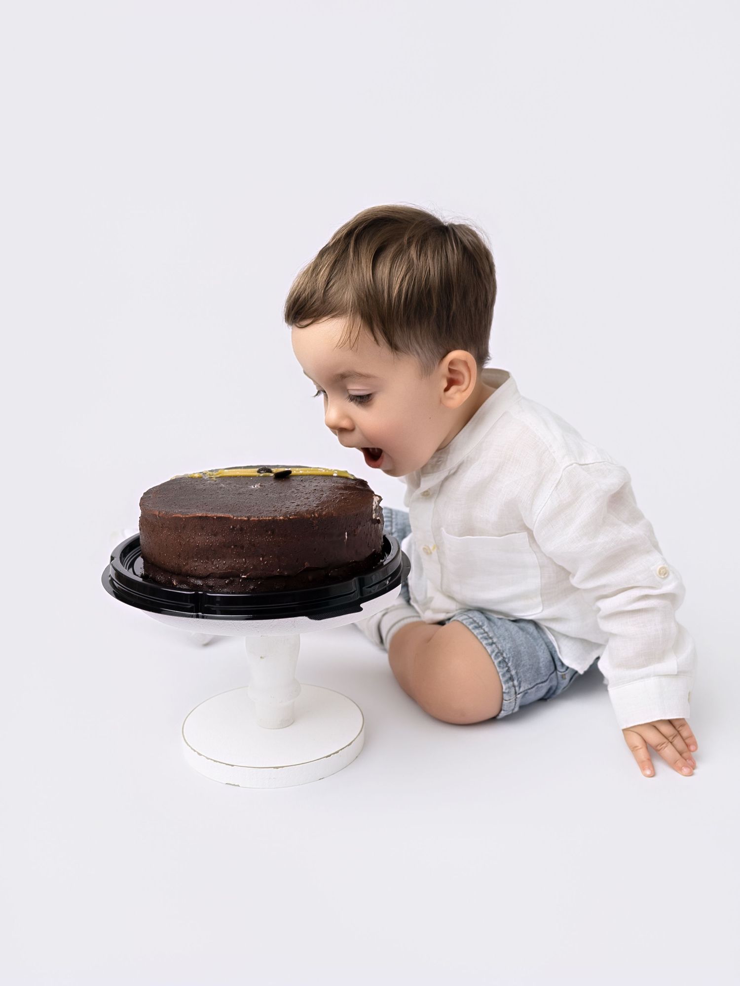 Toddler boy in white shirt and denim shorts leaning toward chocolate birthday cake on white pedestal stand.