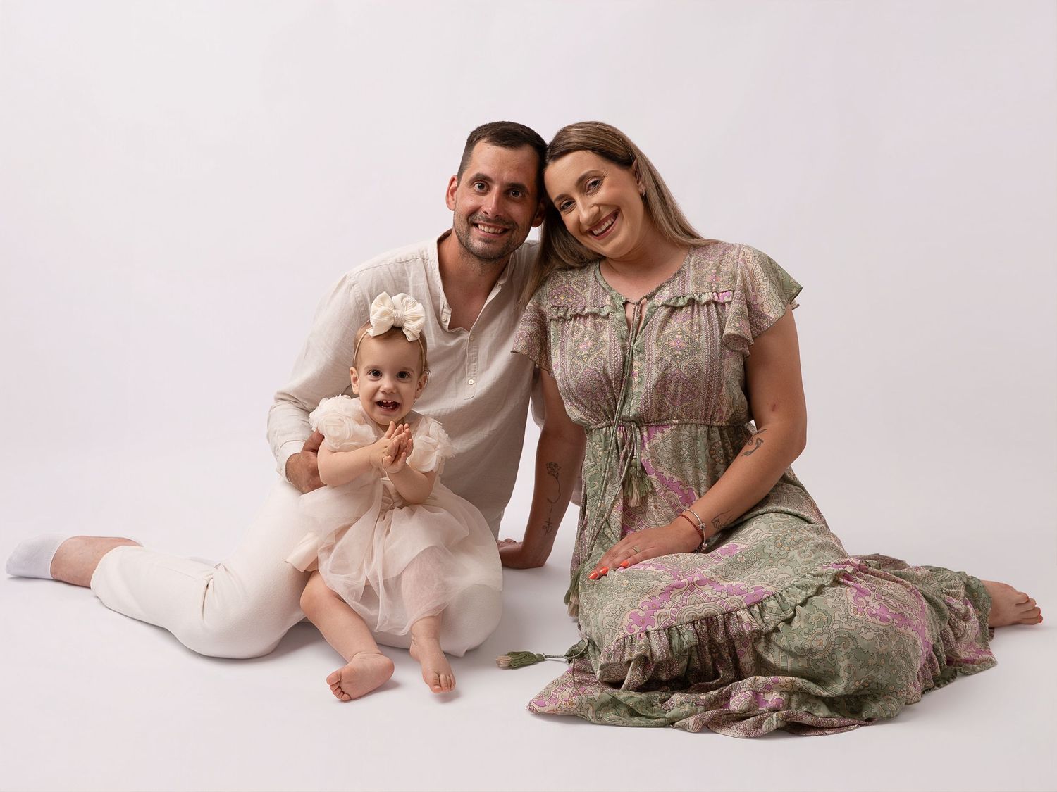 A happy family of three poses for a studio portrait, with parents sitting beside their baby girl in a white dress.