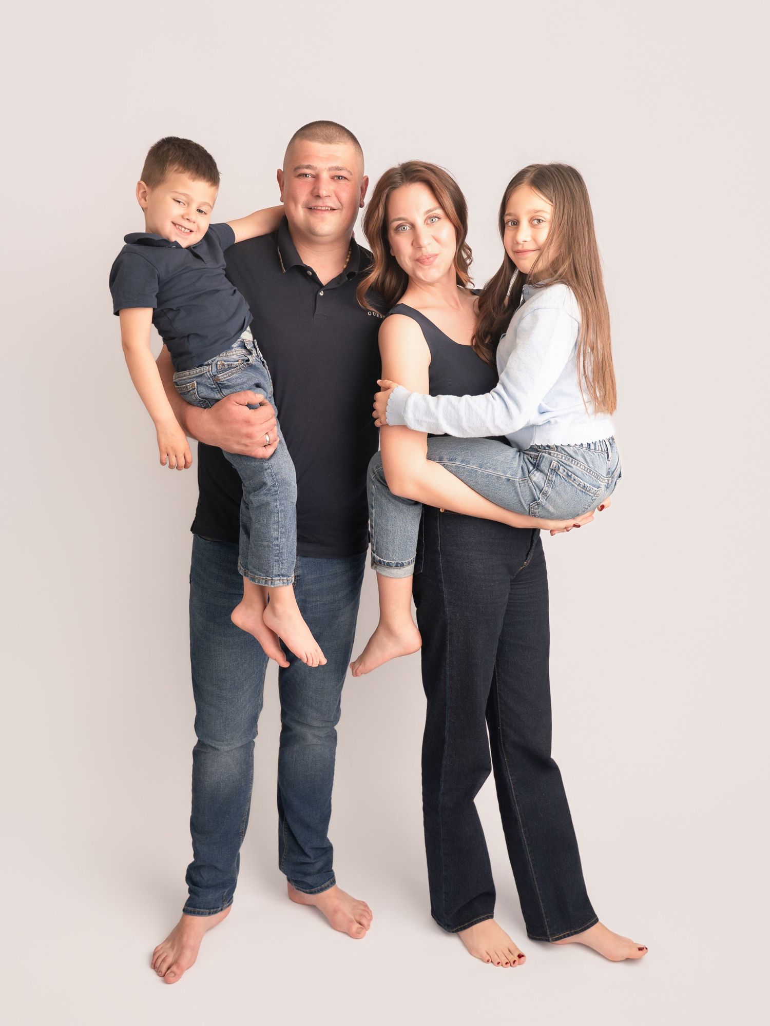 A happy family of four posing barefoot in a studio, parents holding their two young children, dressed in casual dark outfits.
