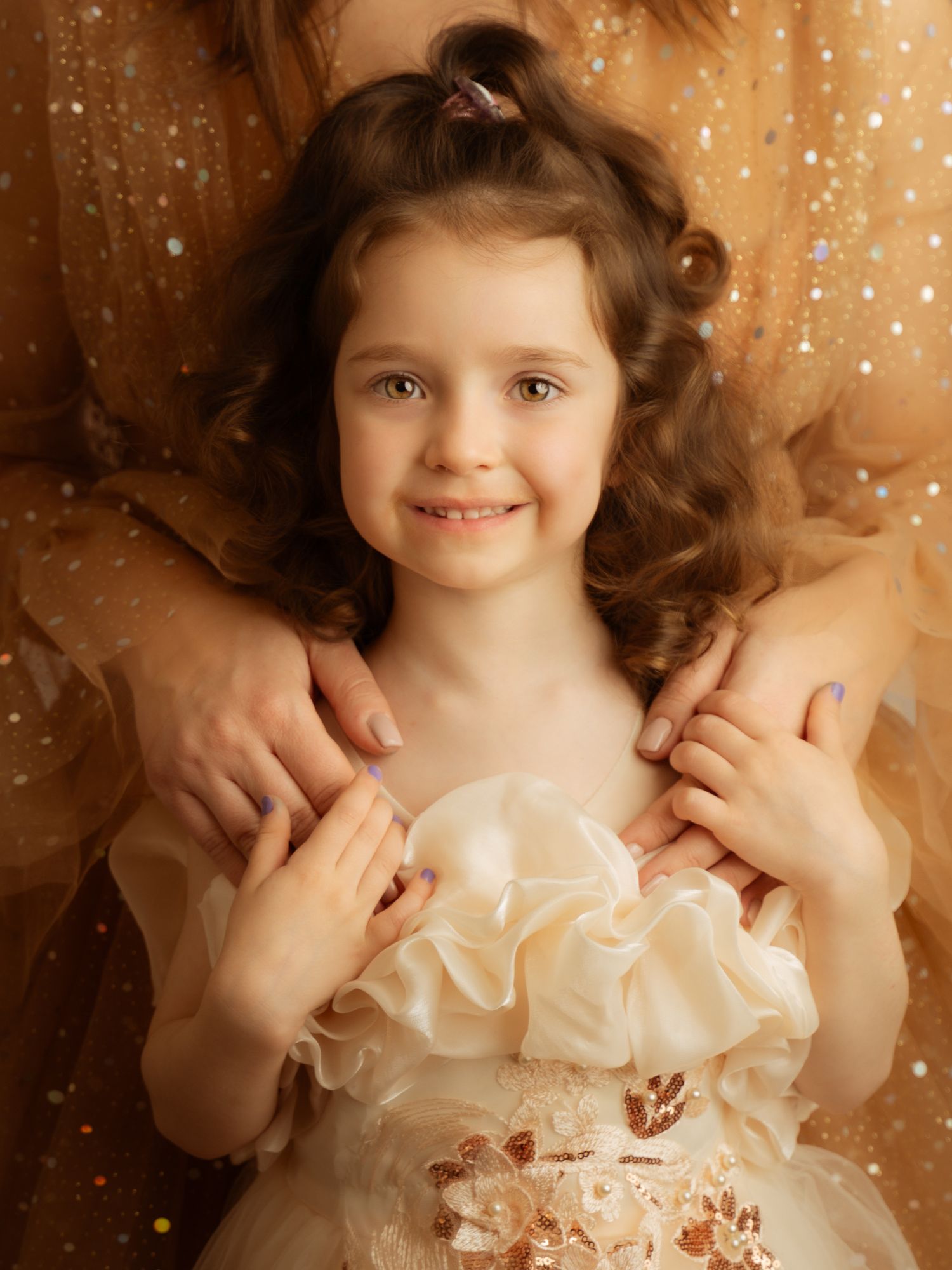 Smiling curly-haired girl in cream ruffled dress held gently by adult hands from behind.