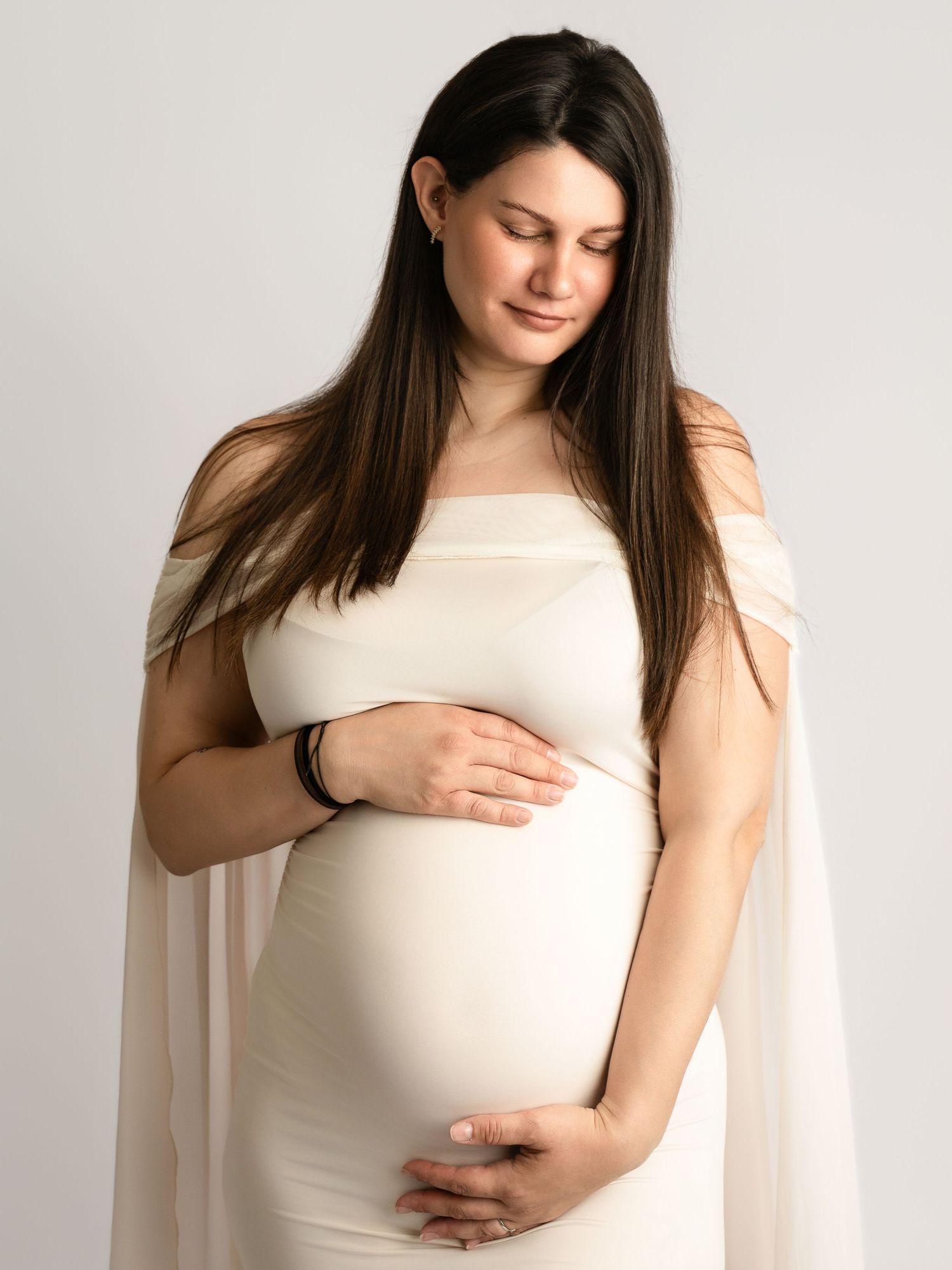 Serene pregnant woman in white cape dress tenderly cradling baby bump, eyes closed.