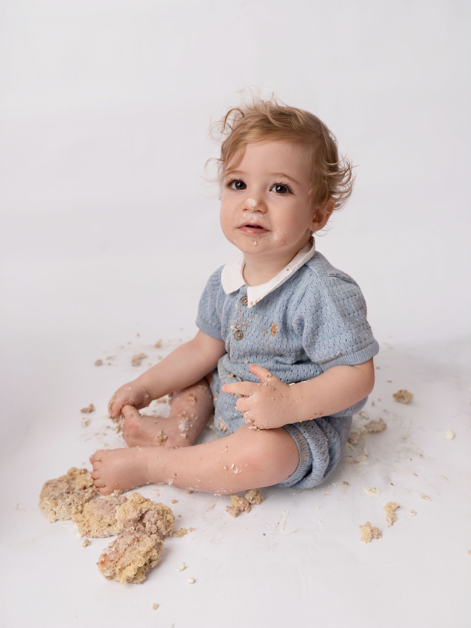 Baby in blue romper sits on white backdrop, covered in cake crumbs during first birthday smash cake photo session.