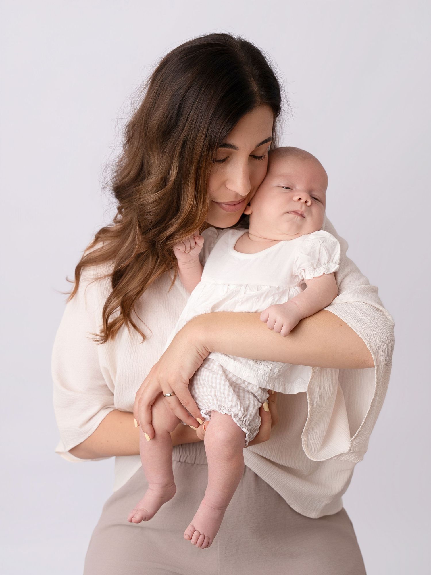 A mother in white tenderly holds her newborn baby against a soft white studio background.
