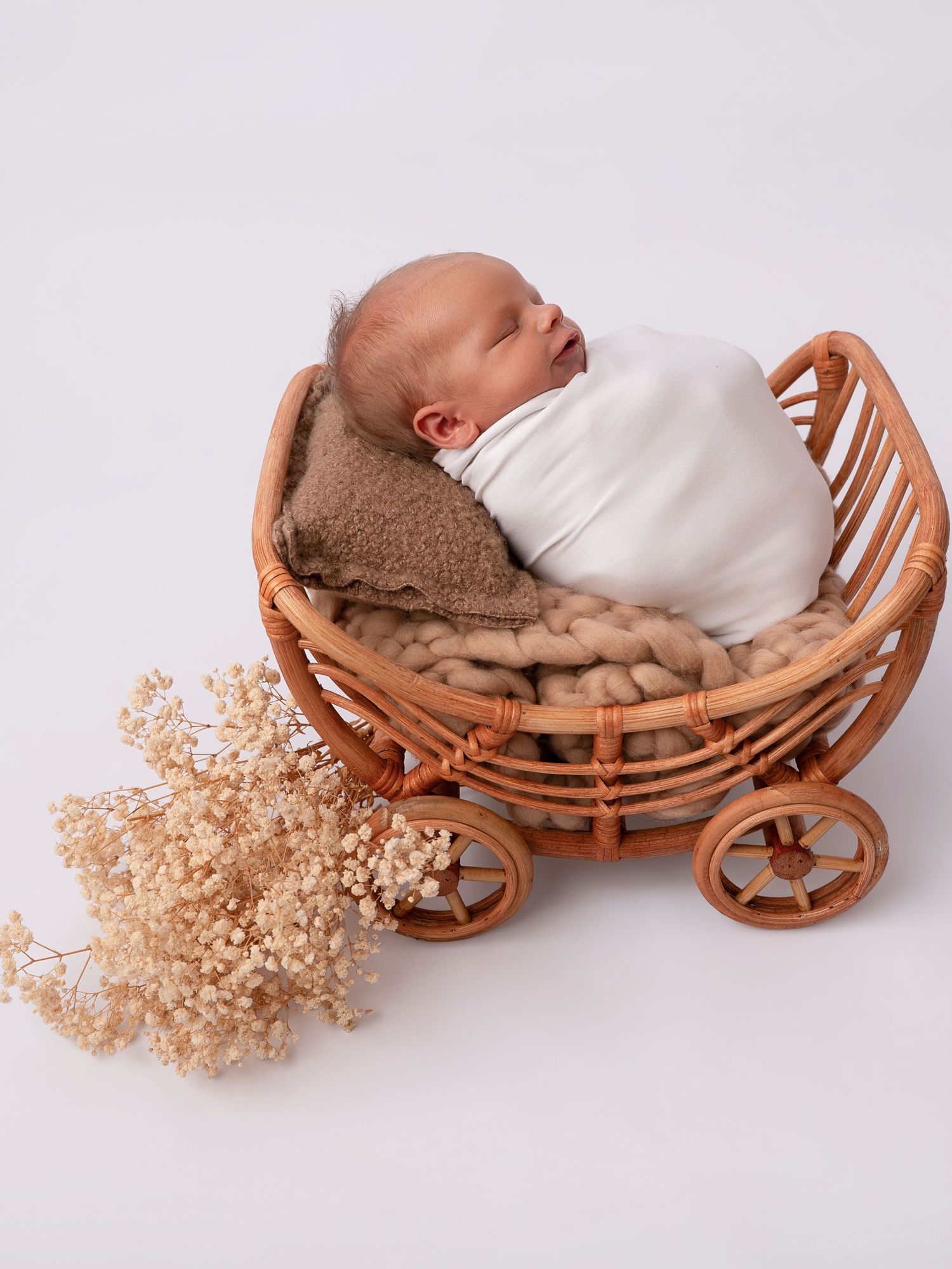 Sleeping newborn baby swaddled in white wrap resting in a rattan pram prop with dried flowers.