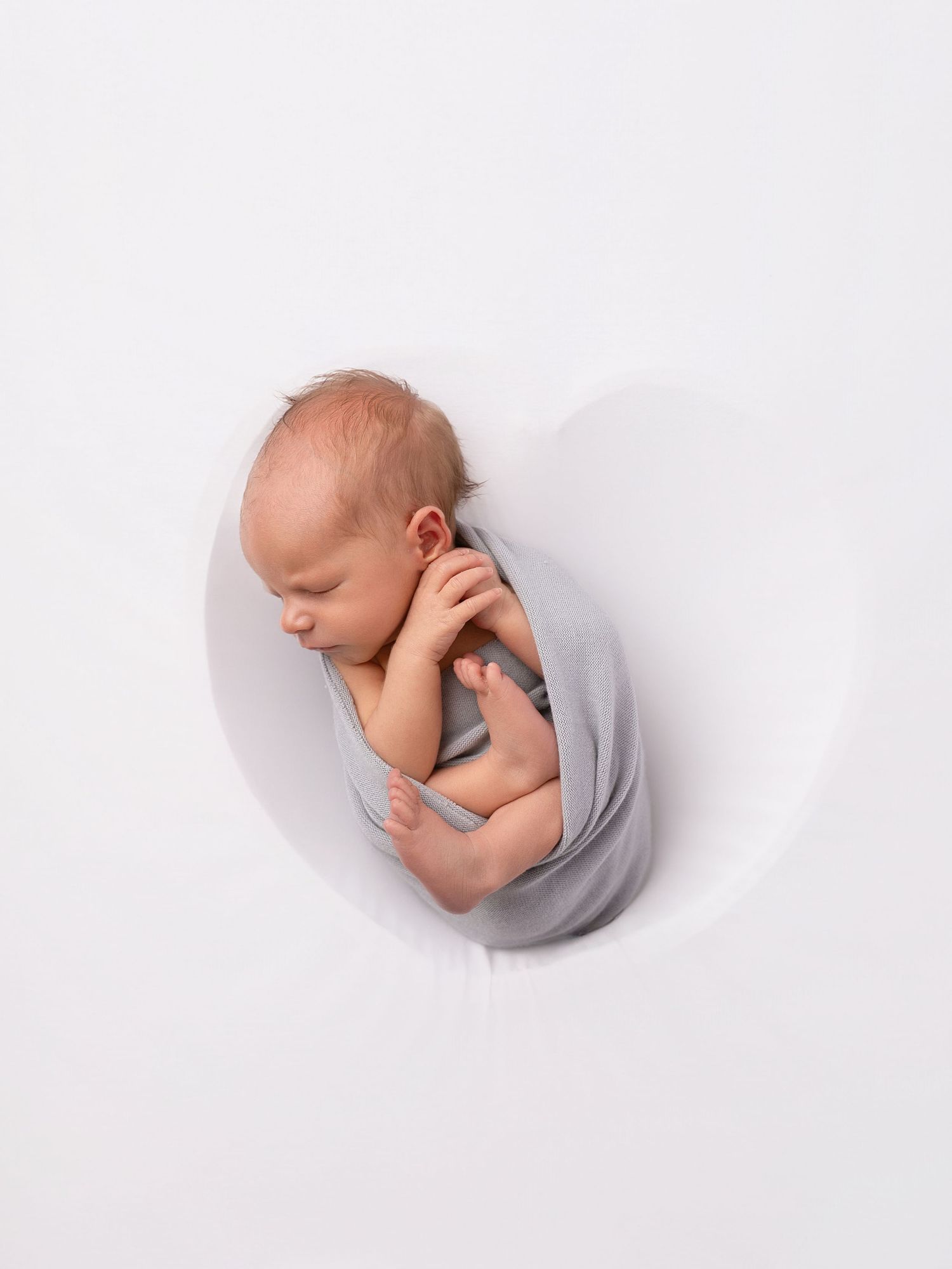 Newborn baby swaddled in gray wrap sleeping peacefully inside a white bowl-shaped prop.