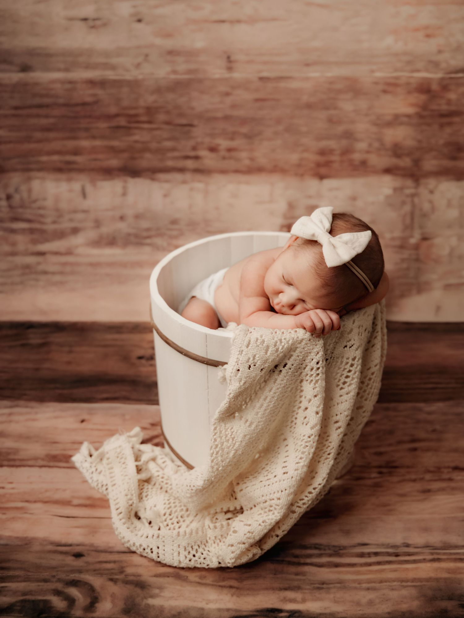 Sleeping newborn baby girl in white bucket with cream knit blanket and bow headband on rustic wood background.
