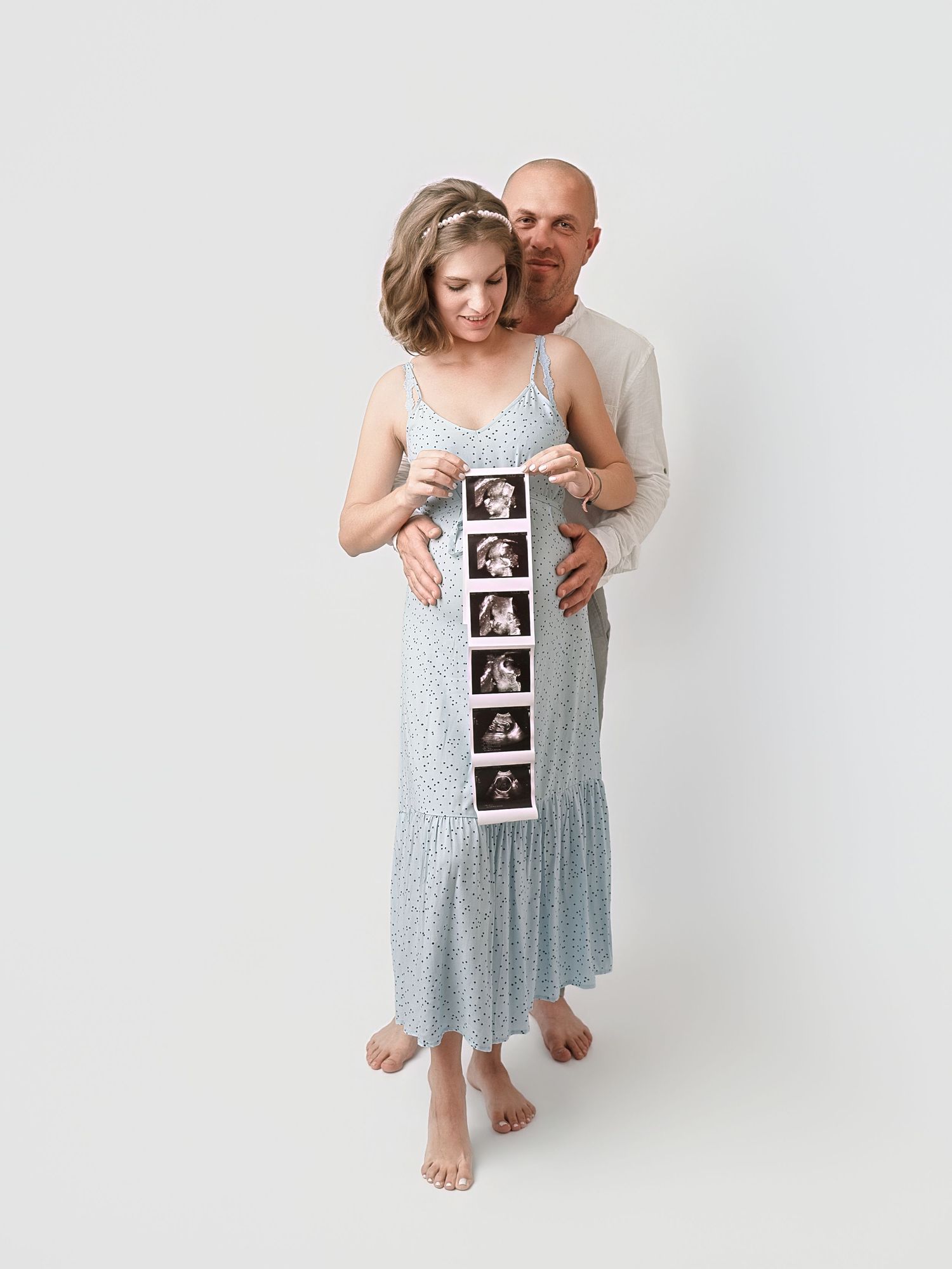 Couple holding ultrasound strip, pregnant woman in blue floral dress embraced by partner on white background.