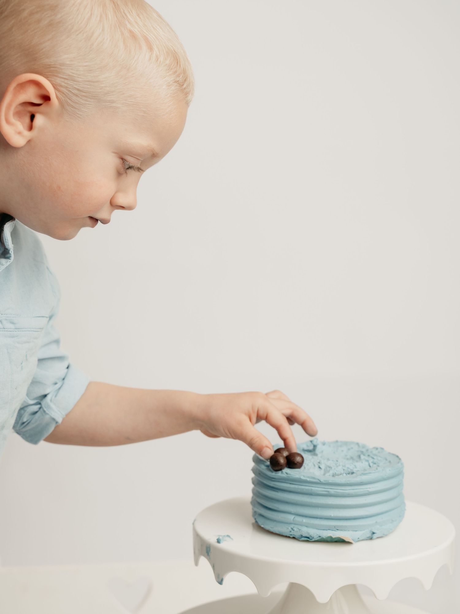 A young blonde toddler in a light blue shirt touches a blue birthday cake on a white cake stand.