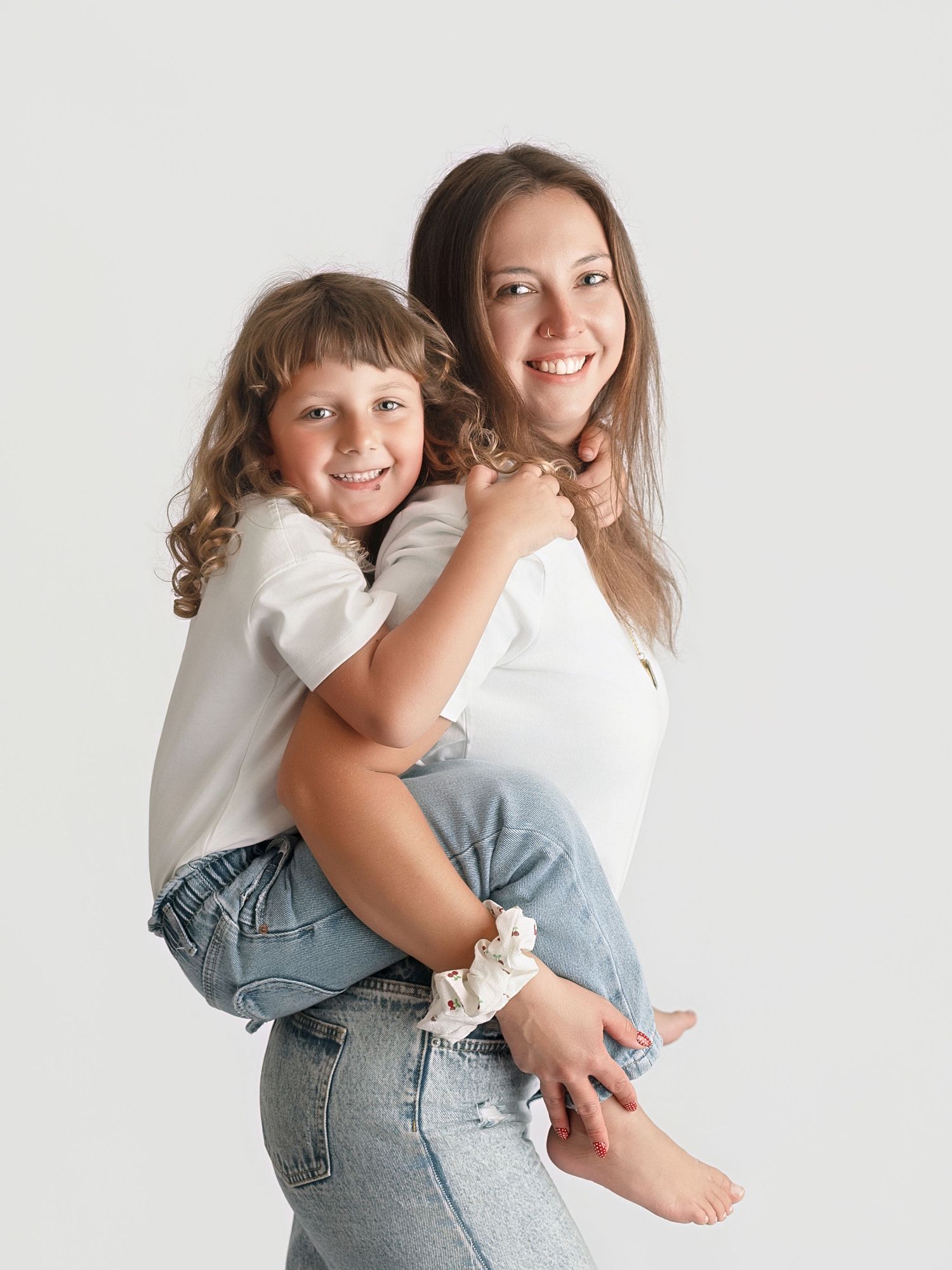 A smiling woman in white t-shirt and jeans gives a happy young girl a piggyback ride against a white background.