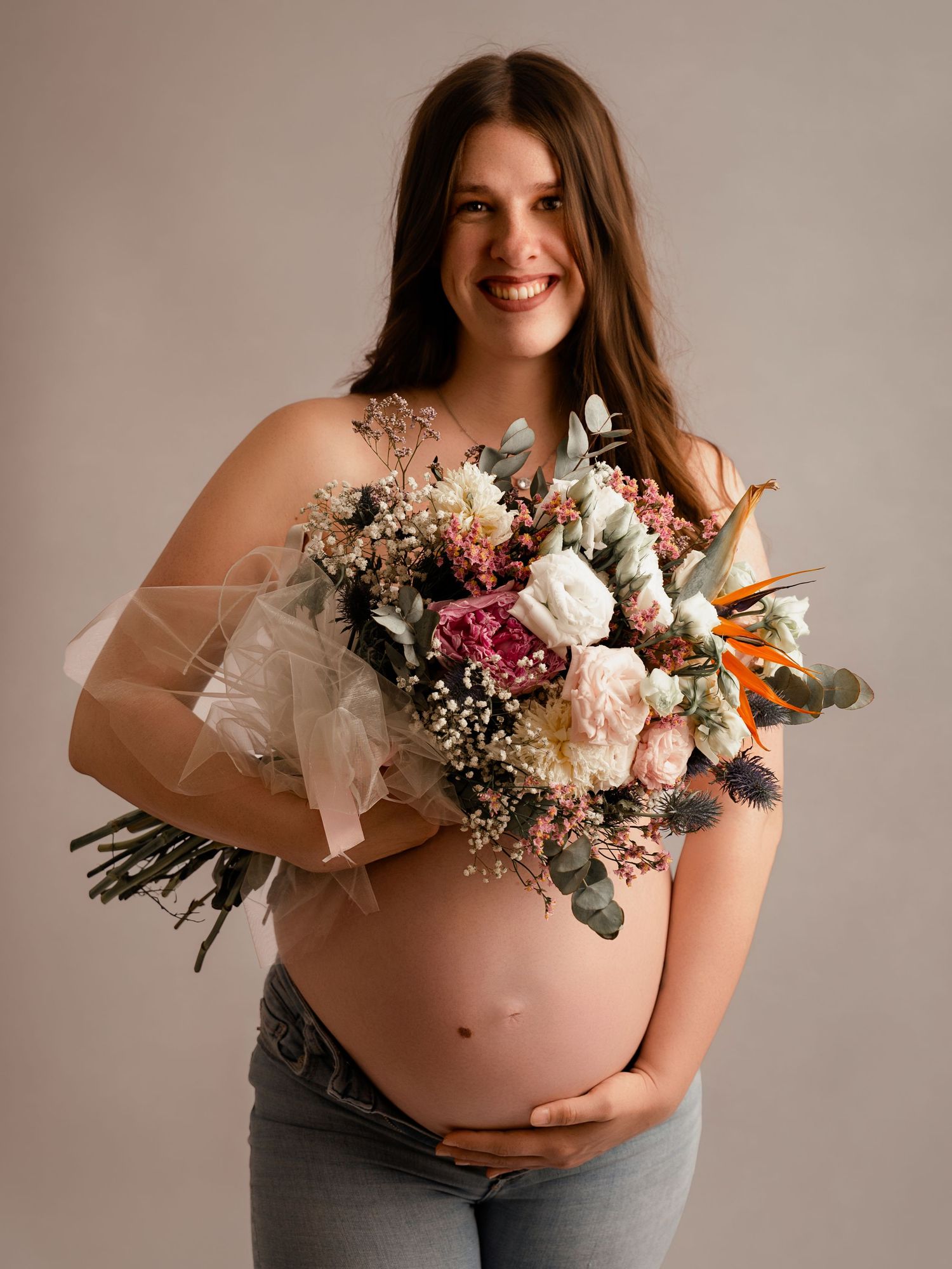 Smiling pregnant woman holding a large bouquet of pink and white flowers while cradling her baby bump in a studio.