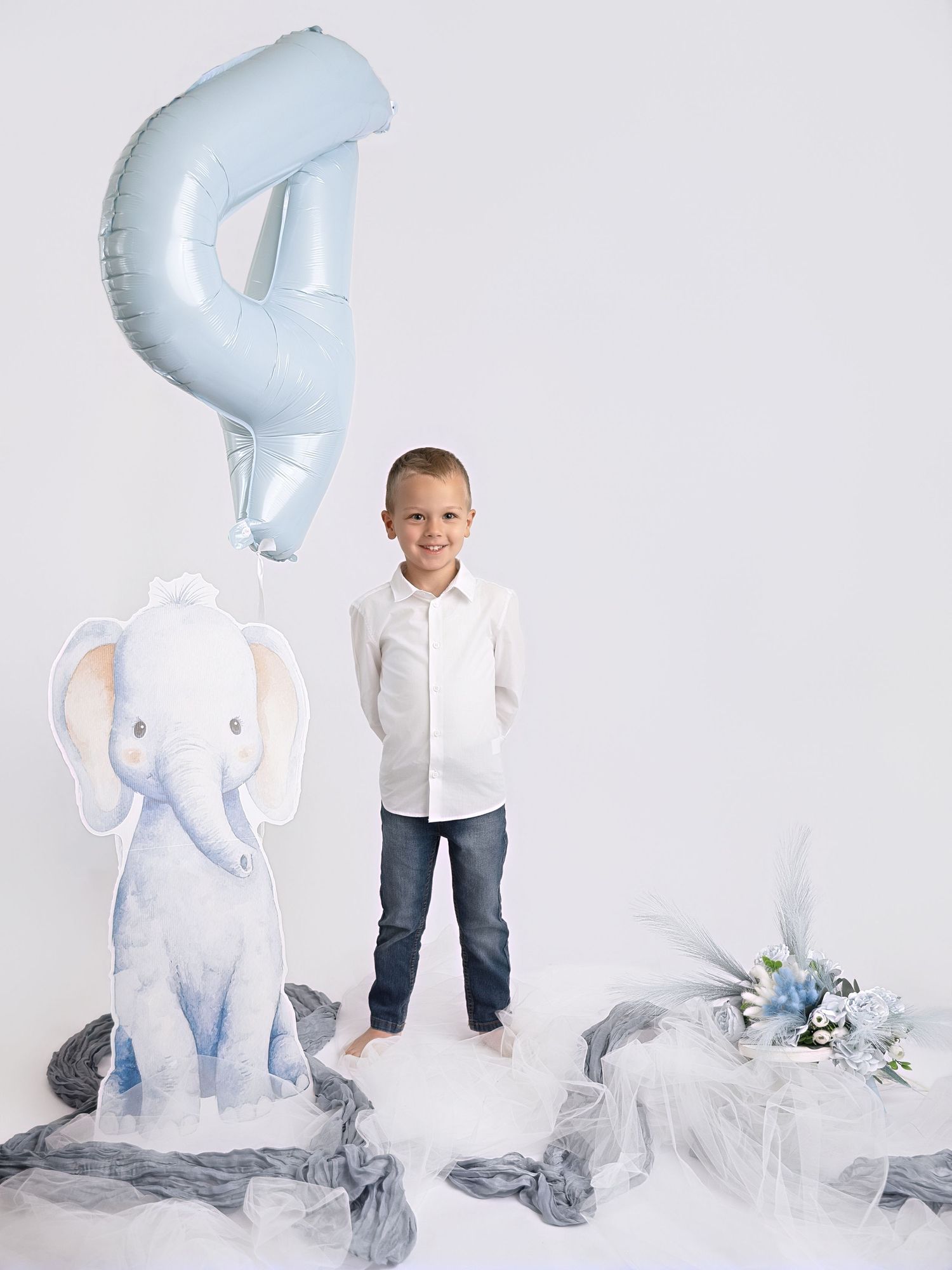 A smiling boy stands beside a cartoon elephant cutout with a blue number 4 balloon for his birthday.