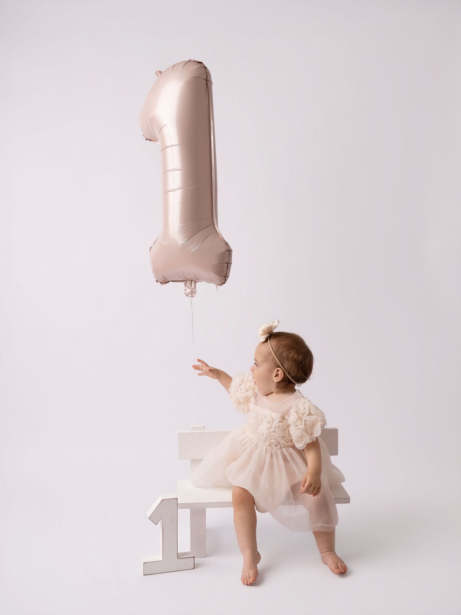 Baby girl in white dress reaching for a rose gold number one balloon at her first birthday photo session.