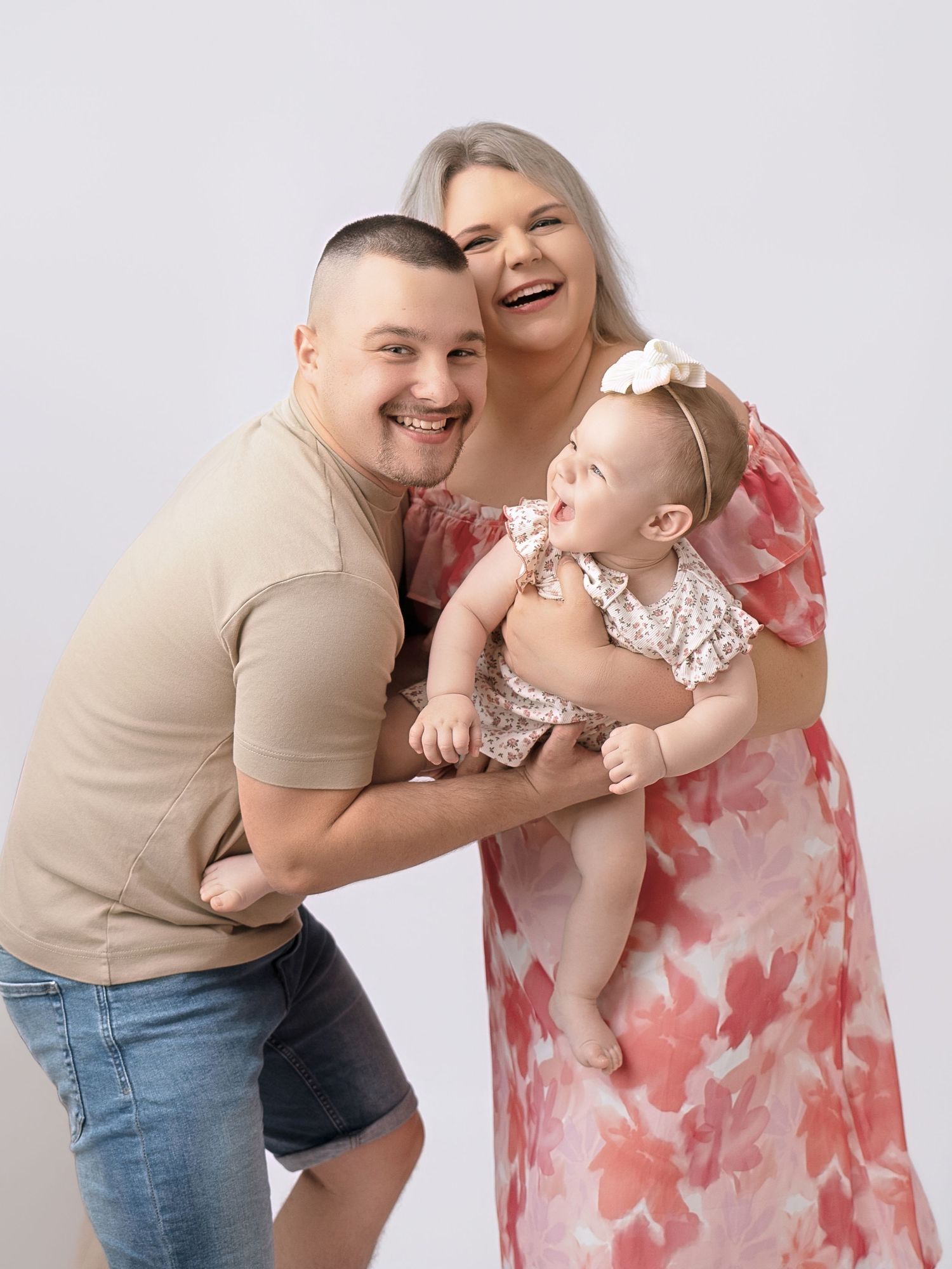 Happy family portrait with smiling parents and adorable baby girl in floral outfits against white background.