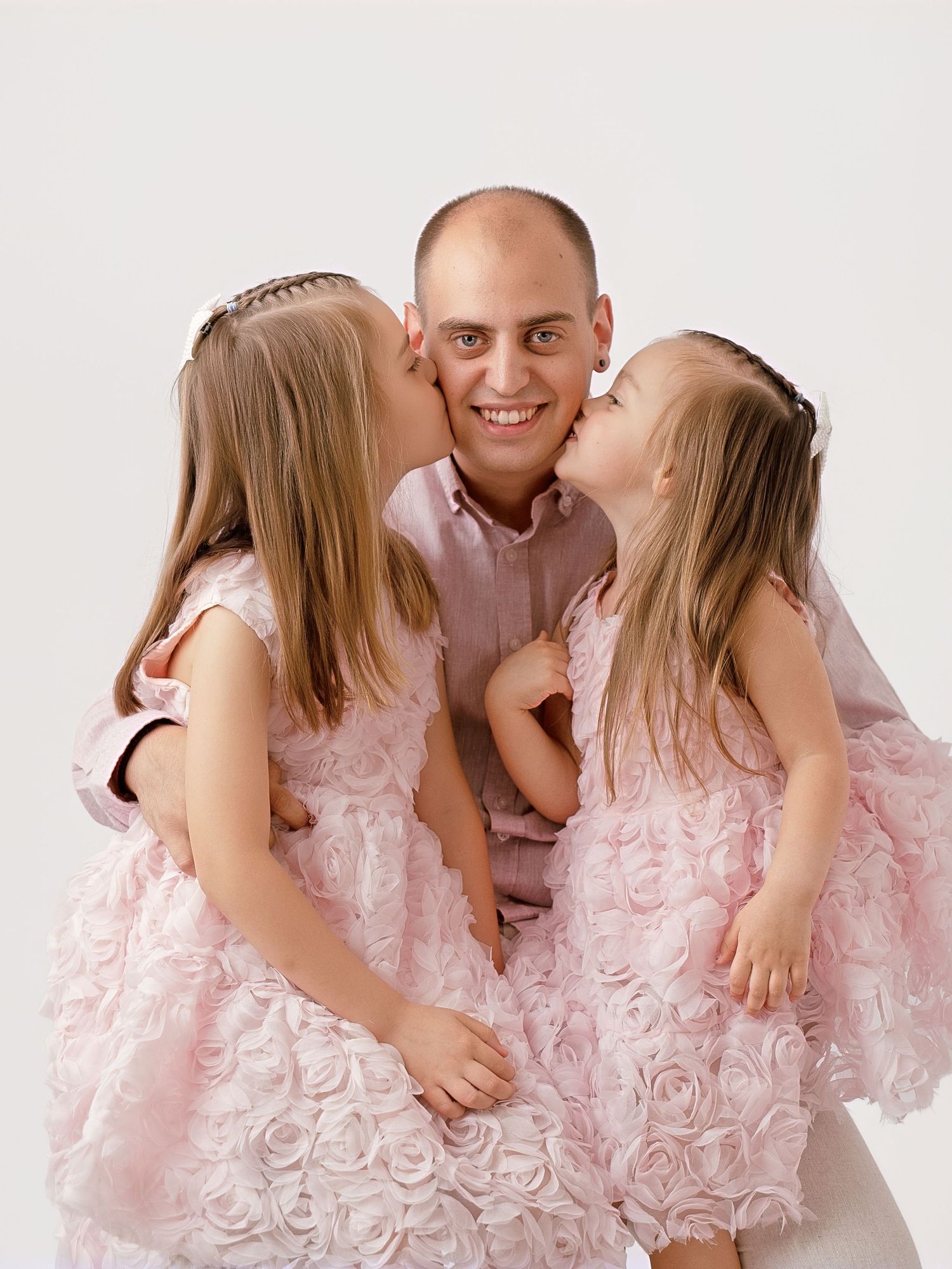 Father smiling as two young daughters in pink dresses give him kisses on the cheeks.
