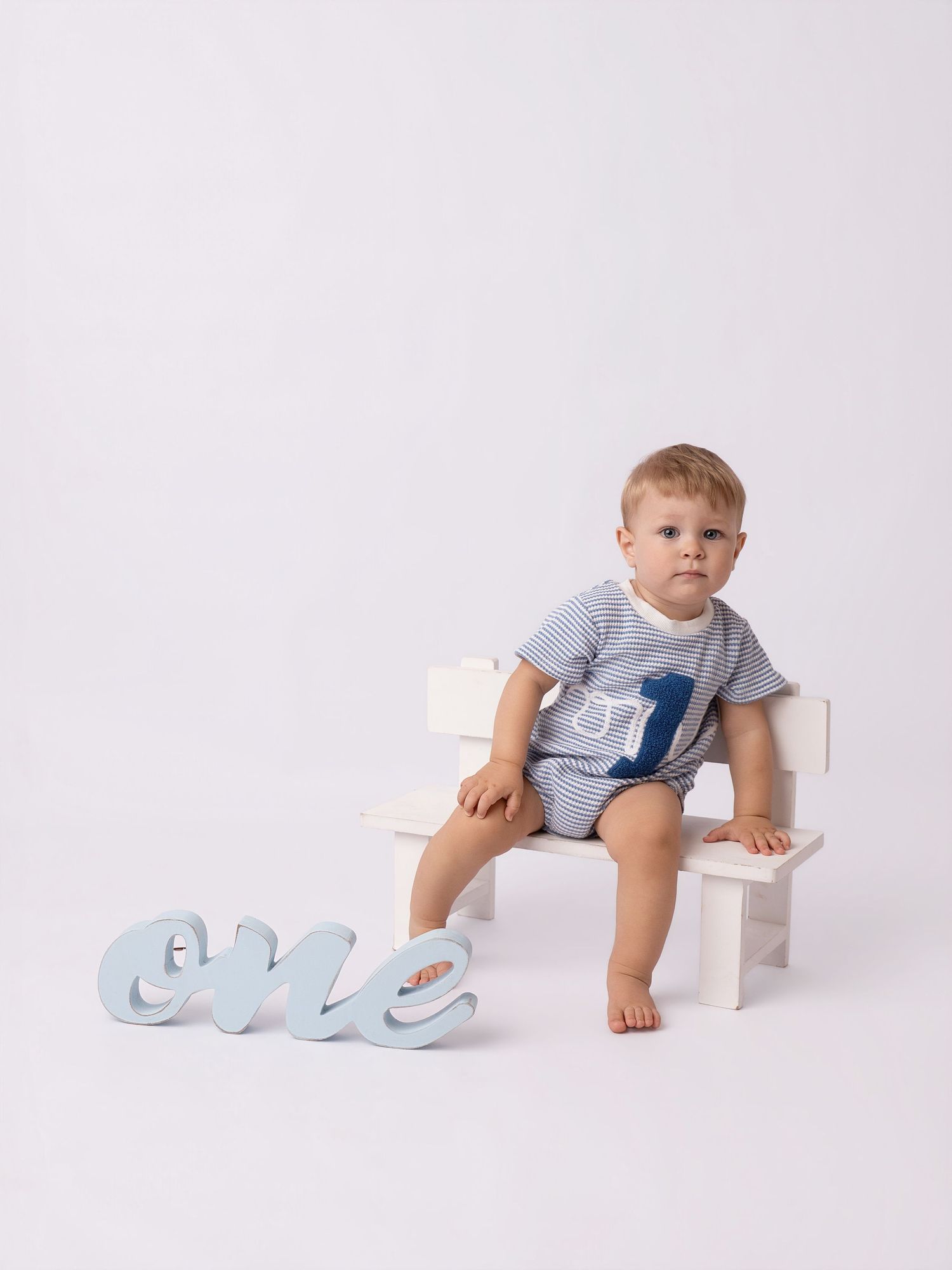 A baby boy sits on a small white bench beside a decorative blue 'one' sign, wearing a striped romper for his first birthday.