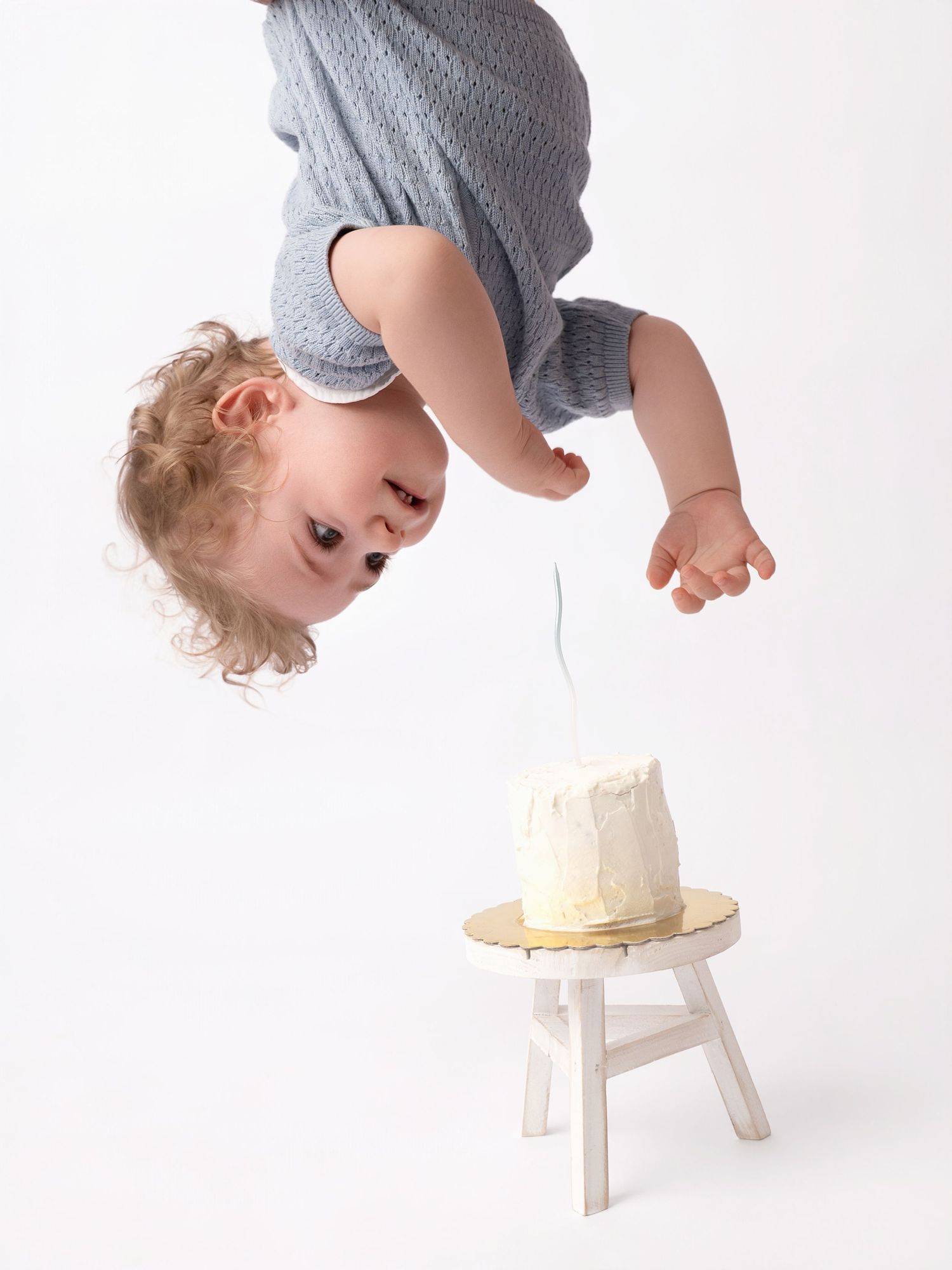 Funny upside down baby reaching toward smash cake on small white stool.