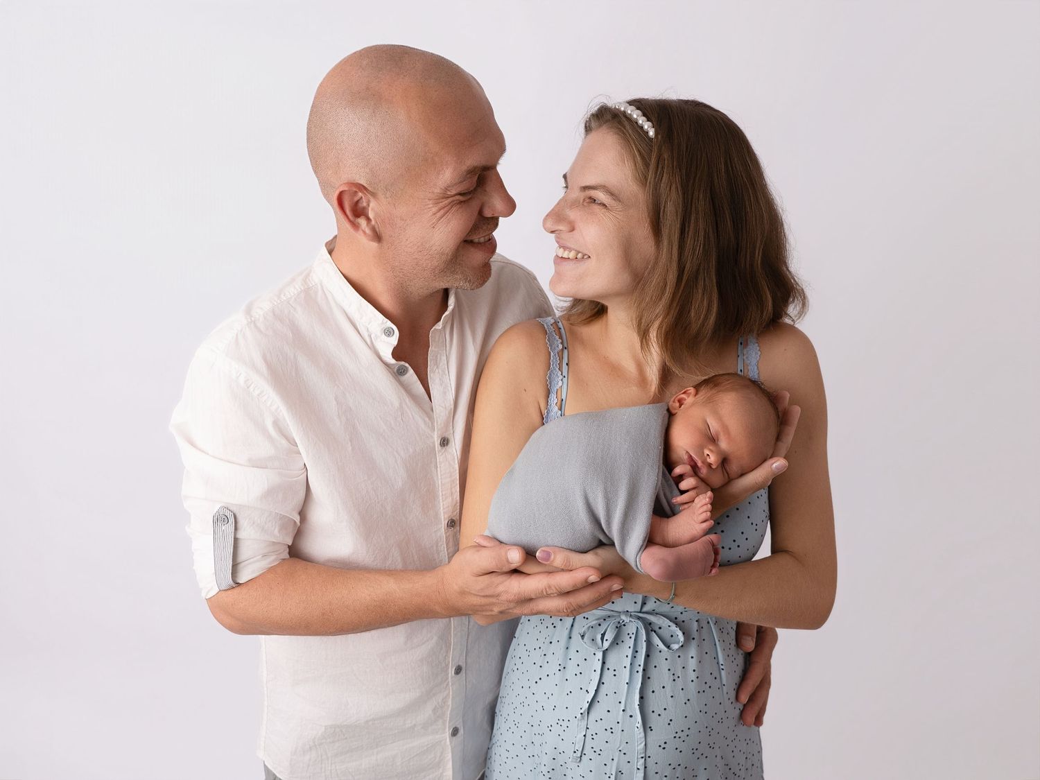 Happy parents smiling at each other while mother holds sleeping newborn baby in a bright studio portrait.