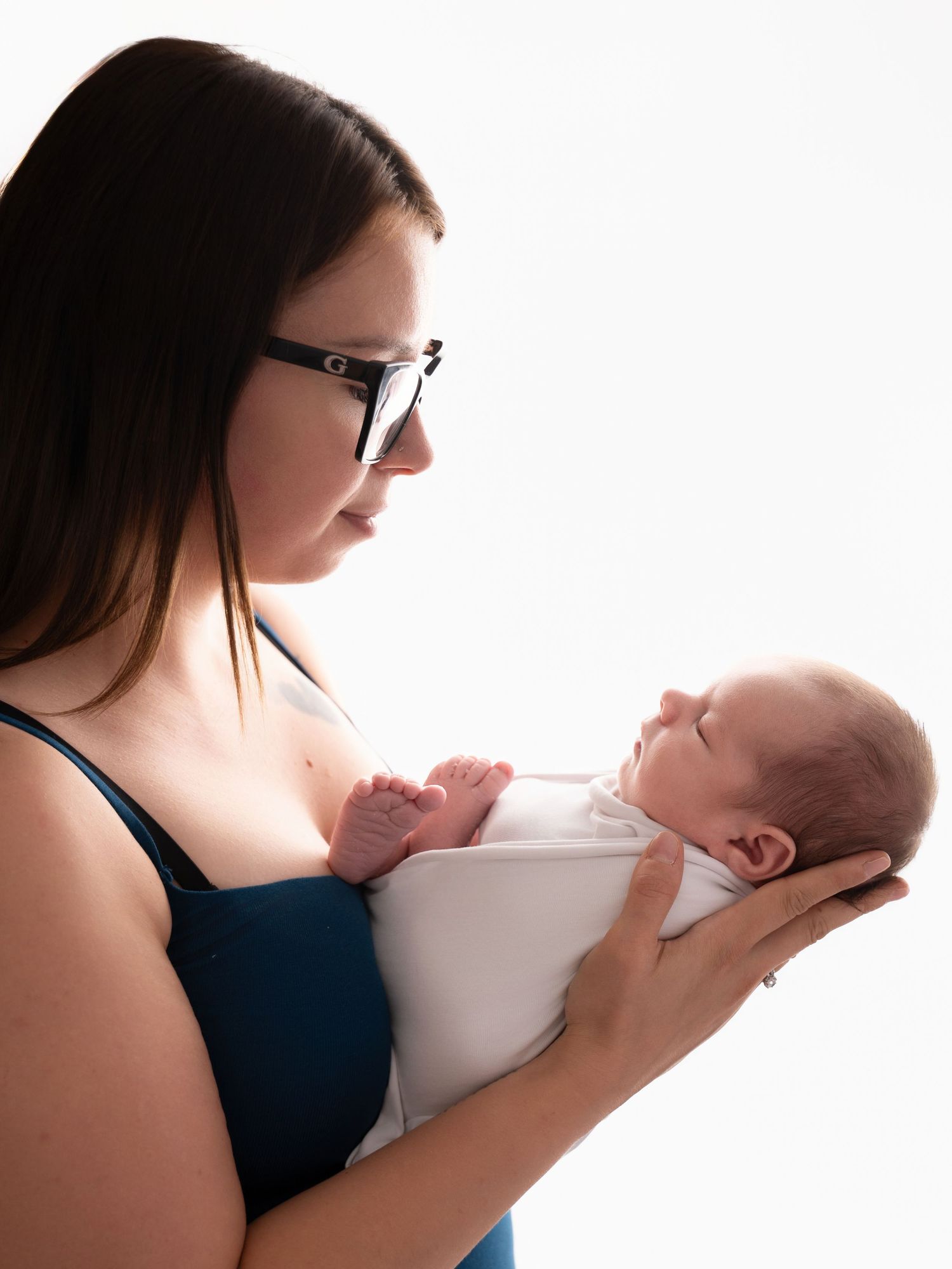A young mother with dark hair and glasses tenderly holds her sleeping newborn baby against a white background.