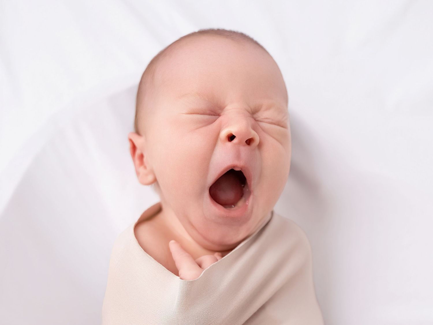 Adorable newborn baby yawning with eyes closed, swaddled in cream wrap on white background.