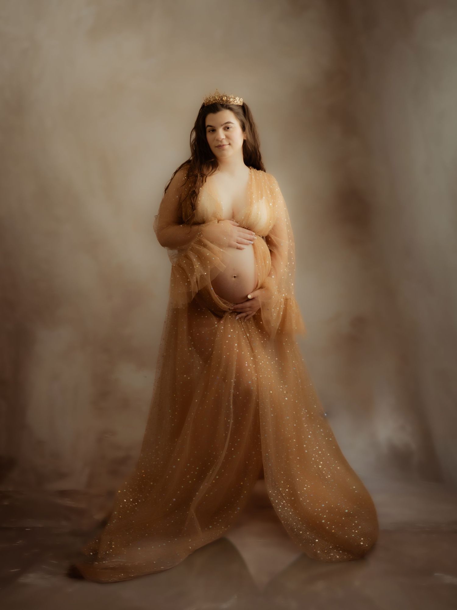 Pregnant woman wearing a gold glitter tulle maternity gown and crown cradling her baby bump in a studio.