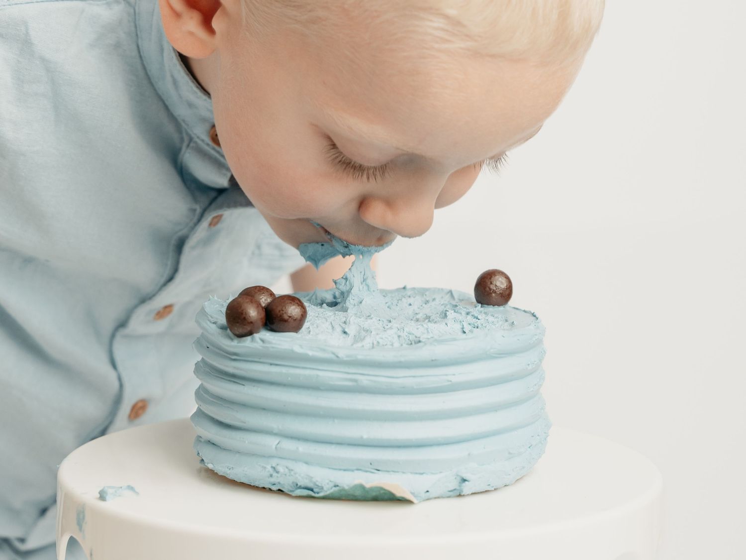 A young toddler in a light blue shirt leans in to eat a blue frosted birthday cake on a white stand.