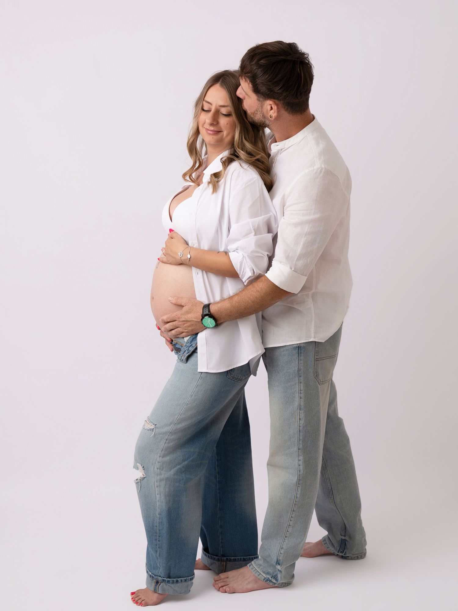 Pregnant couple in white shirts and jeans posing together, man embracing woman from behind in studio.
