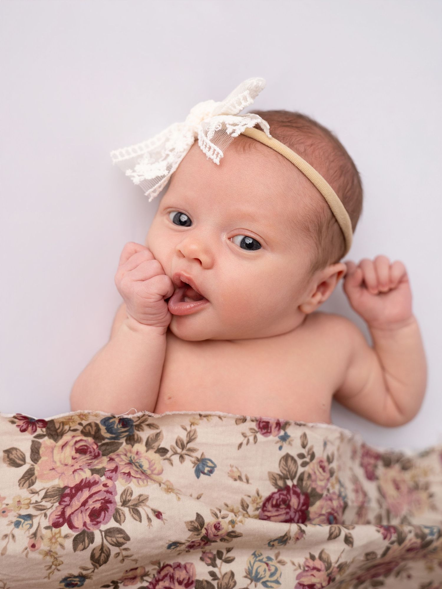 Newborn baby girl with blue eyes wearing a lace bow headband, wrapped in a floral blanket during a studio photo session.