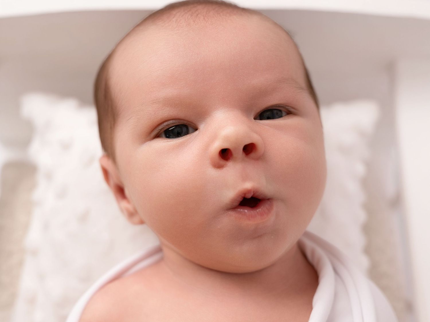 Close-up portrait of a wide-eyed newborn baby wrapped in white, looking up with a curious expression.