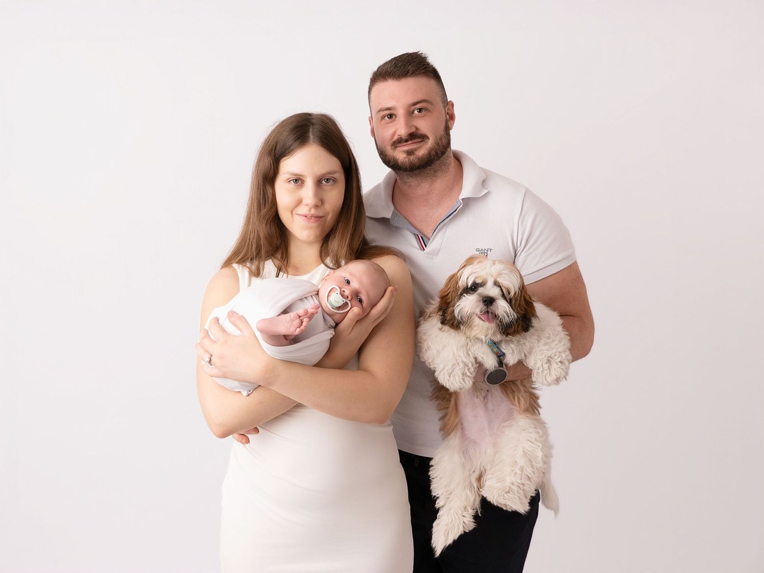 A young family poses for a newborn portrait, with the mother holding a baby and the father holding a Shih Tzu dog.