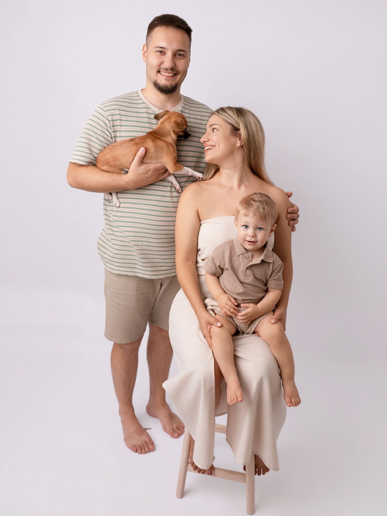 Young family portrait with man, woman, toddler, and small dog posing together in a bright studio setting.