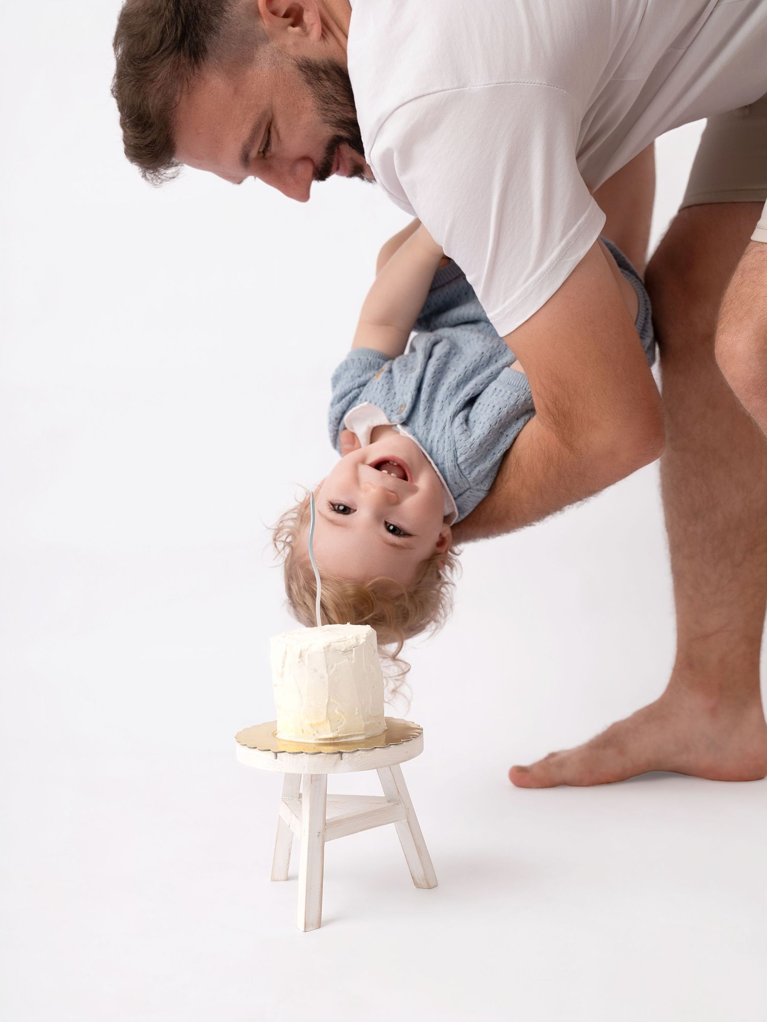 Father playfully holds baby upside down near a small white birthday cake on a stool against white background.