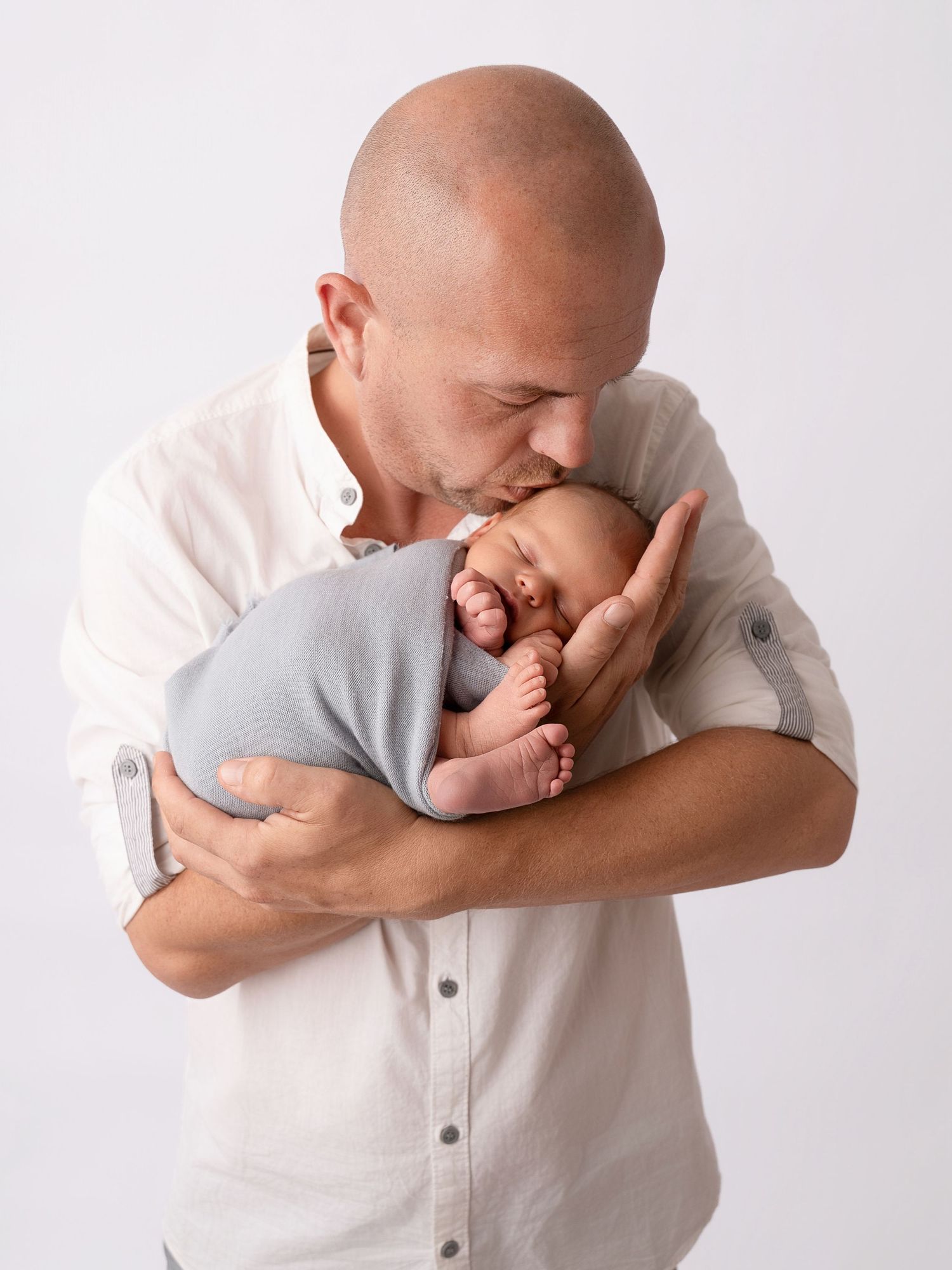 Bald father in white shirt tenderly kissing his swaddled newborn baby wrapped in a gray cloth.